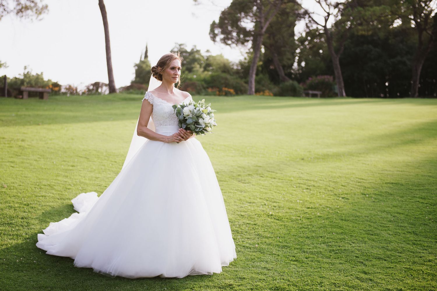 Bride in flowing white gown poses on expansive green lawn with white bouquet.