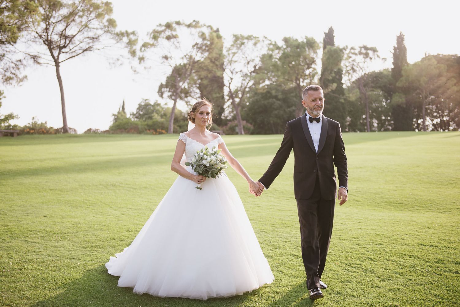Newlywed couple walking hand in hand across sunny green lawn in formal wedding attire.