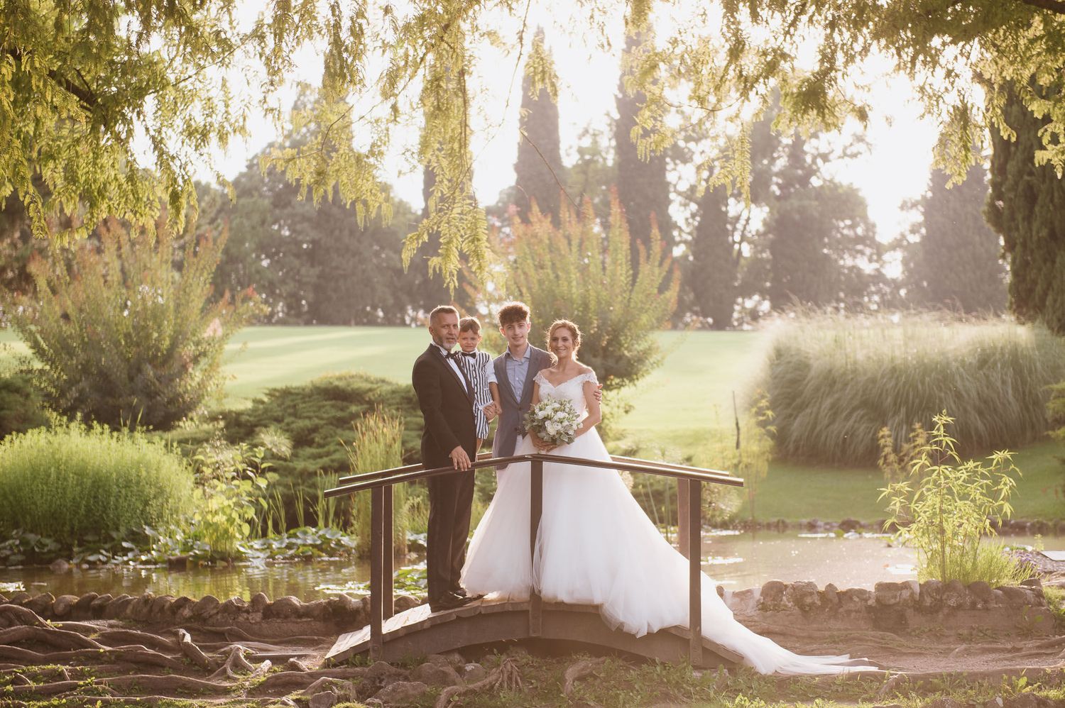 Wedding party poses on rustic wooden bridge in sunlit garden setting.