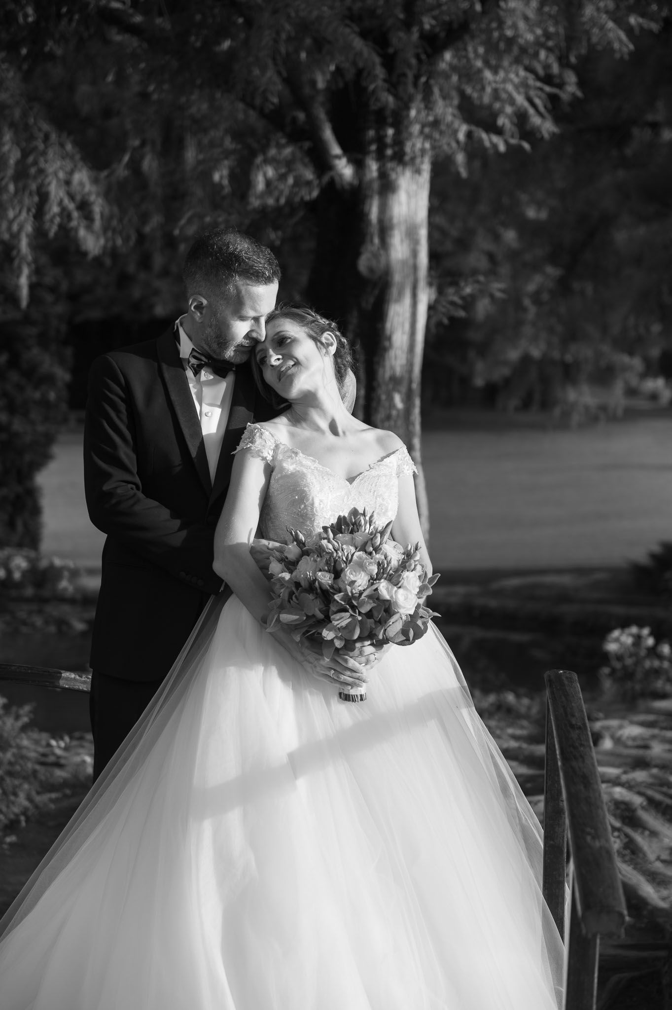 A romantic black and white wedding portrait of a couple embracing outdoors with the bride holding a floral bouquet.