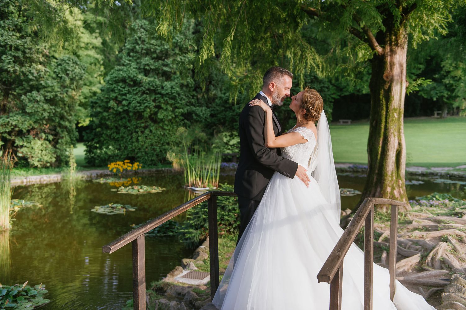 A newlywed couple shares an intimate moment on a wooden bridge over a pond in a garden setting.