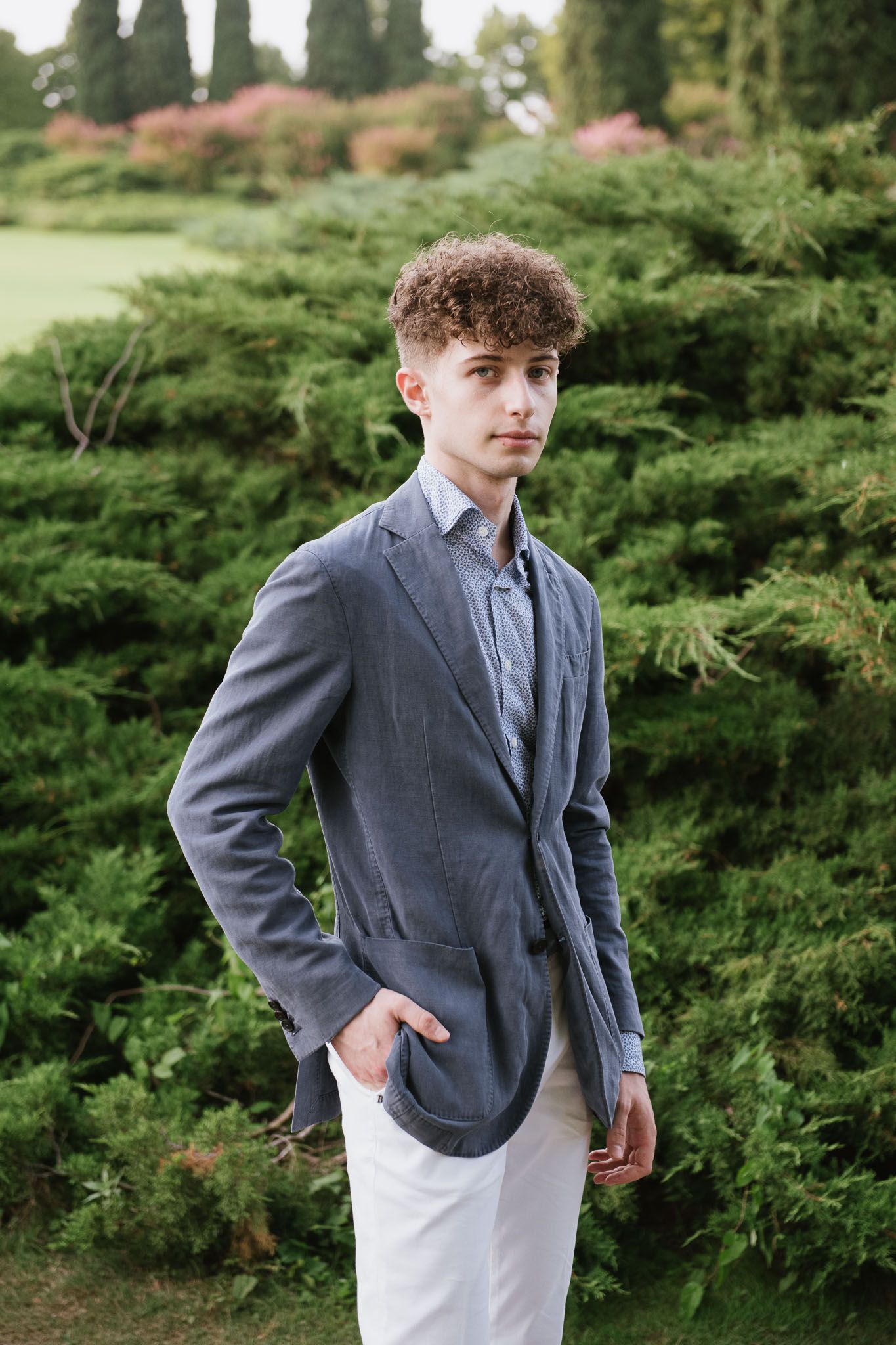 A young man in a gray blazer and white pants poses against evergreen foliage in a garden.