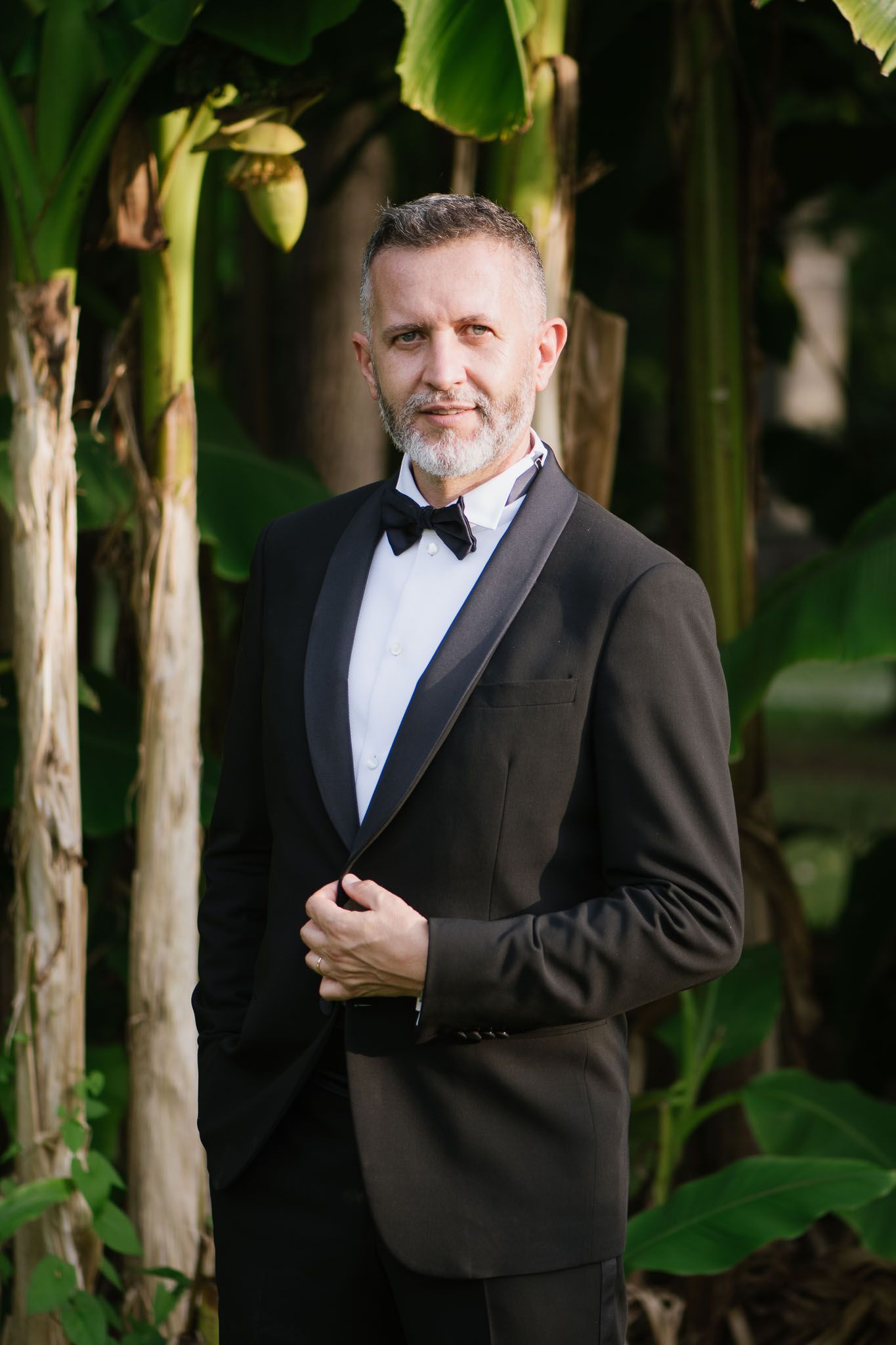 A formal portrait of a gentleman in a classic black tuxedo standing among tropical plants.