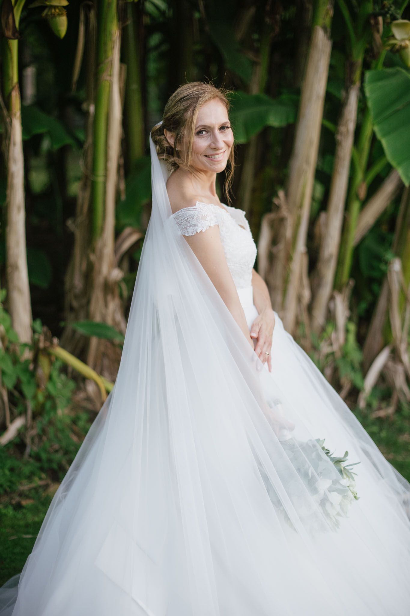 A bride in a flowing white gown and veil poses among tropical foliage in natural lighting.