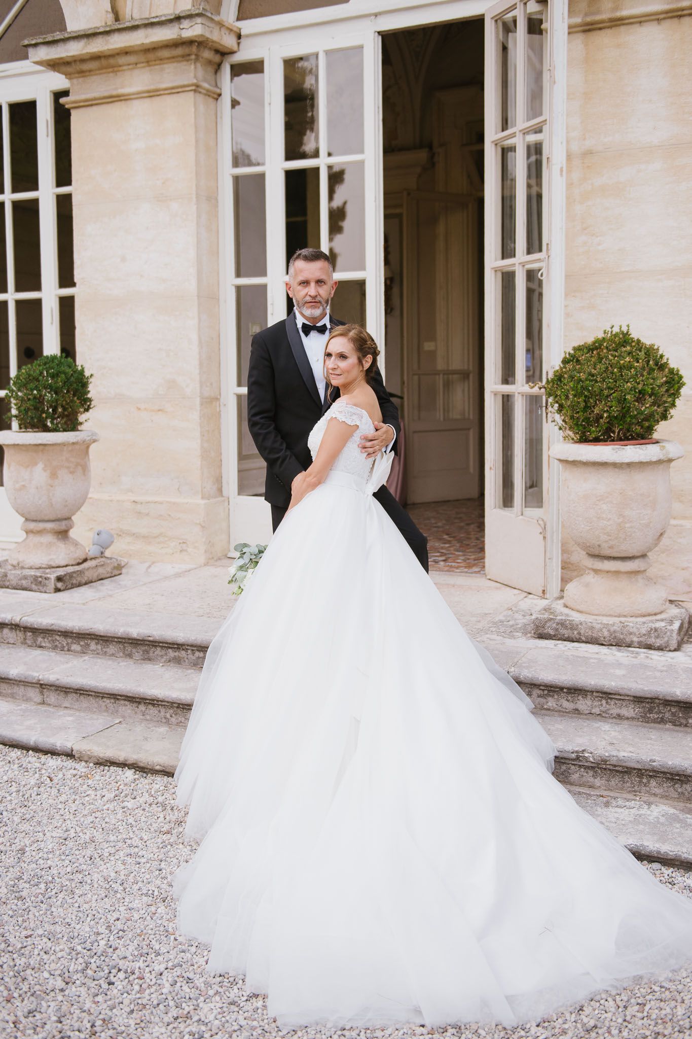 A romantic wedding couple poses in front of an elegant stone mansion with French doors and decorative urns.