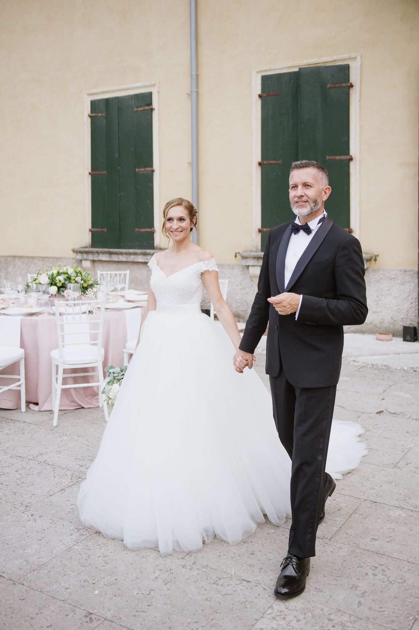 A joyful wedding couple walking hand in hand past outdoor reception tables with green shuttered windows.