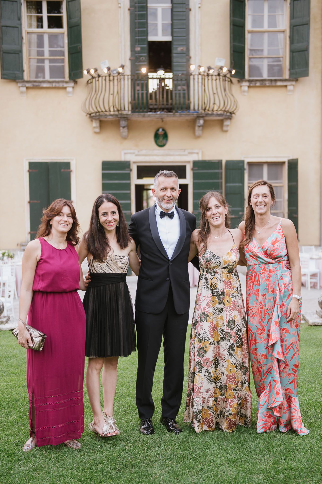 A formal group photo of wedding guests in colorful dresses on a manicured lawn in front of a villa.