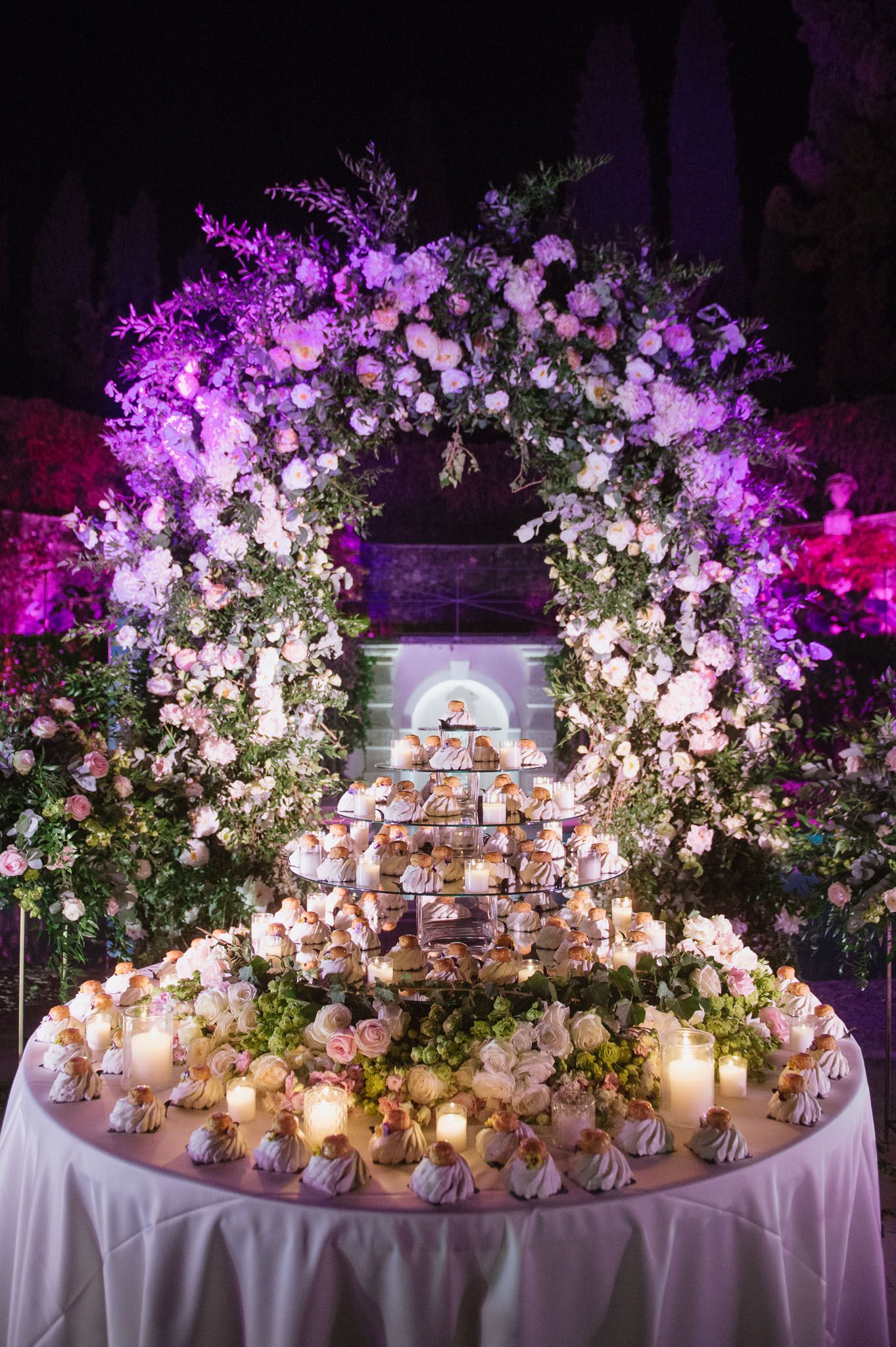 An illuminated floral arch with purple lighting frames a tiered display of treats and candles.
