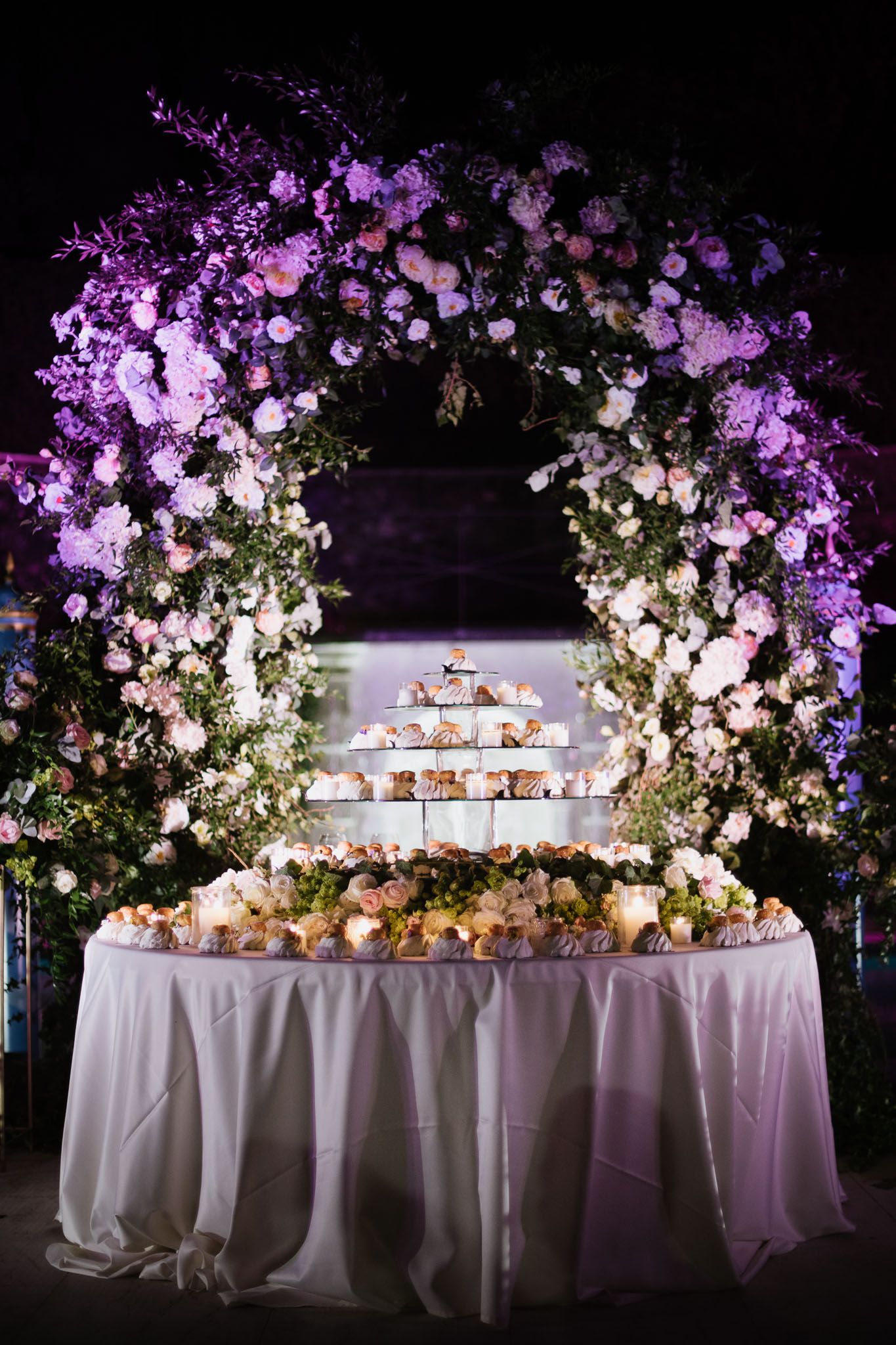 A stunning floral arch with purple lighting frames a tiered dessert display on an elegant white tablecloth.