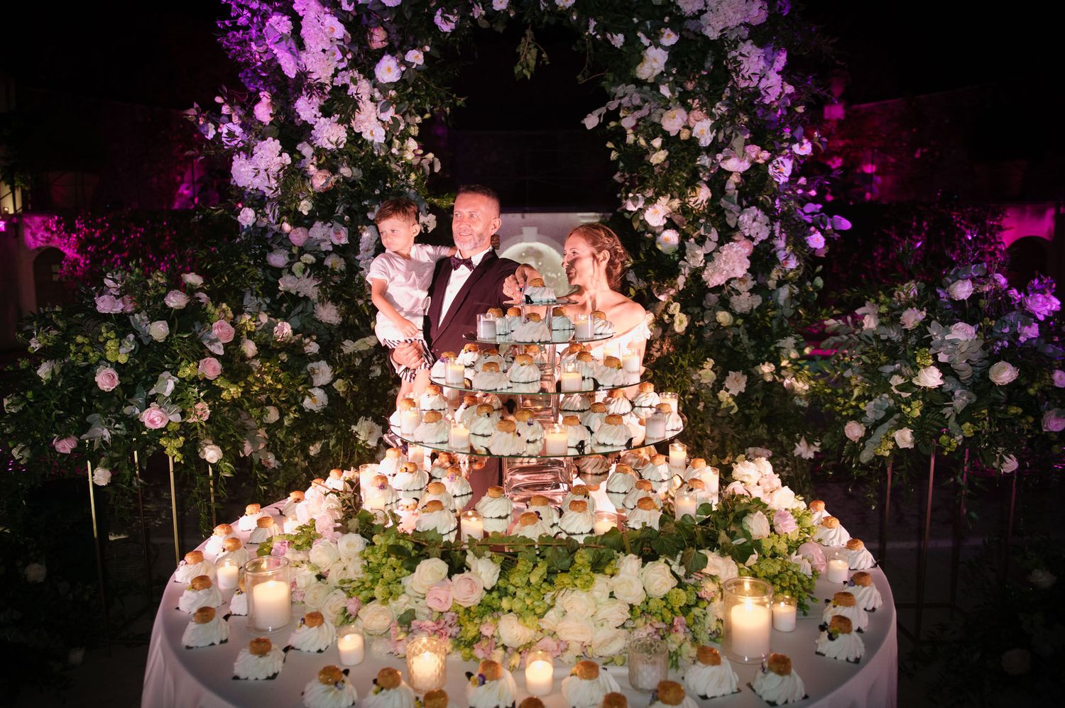 A tiered dessert display illuminated by candlelight under a floral arch with purple accent lighting.