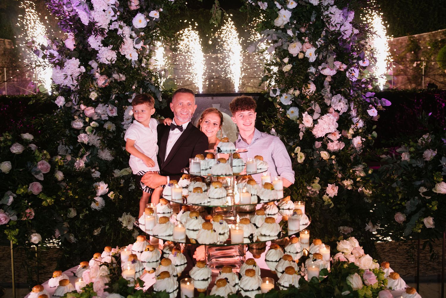 A dessert display surrounded by a floral arch with purple lighting and sparklers in the background.