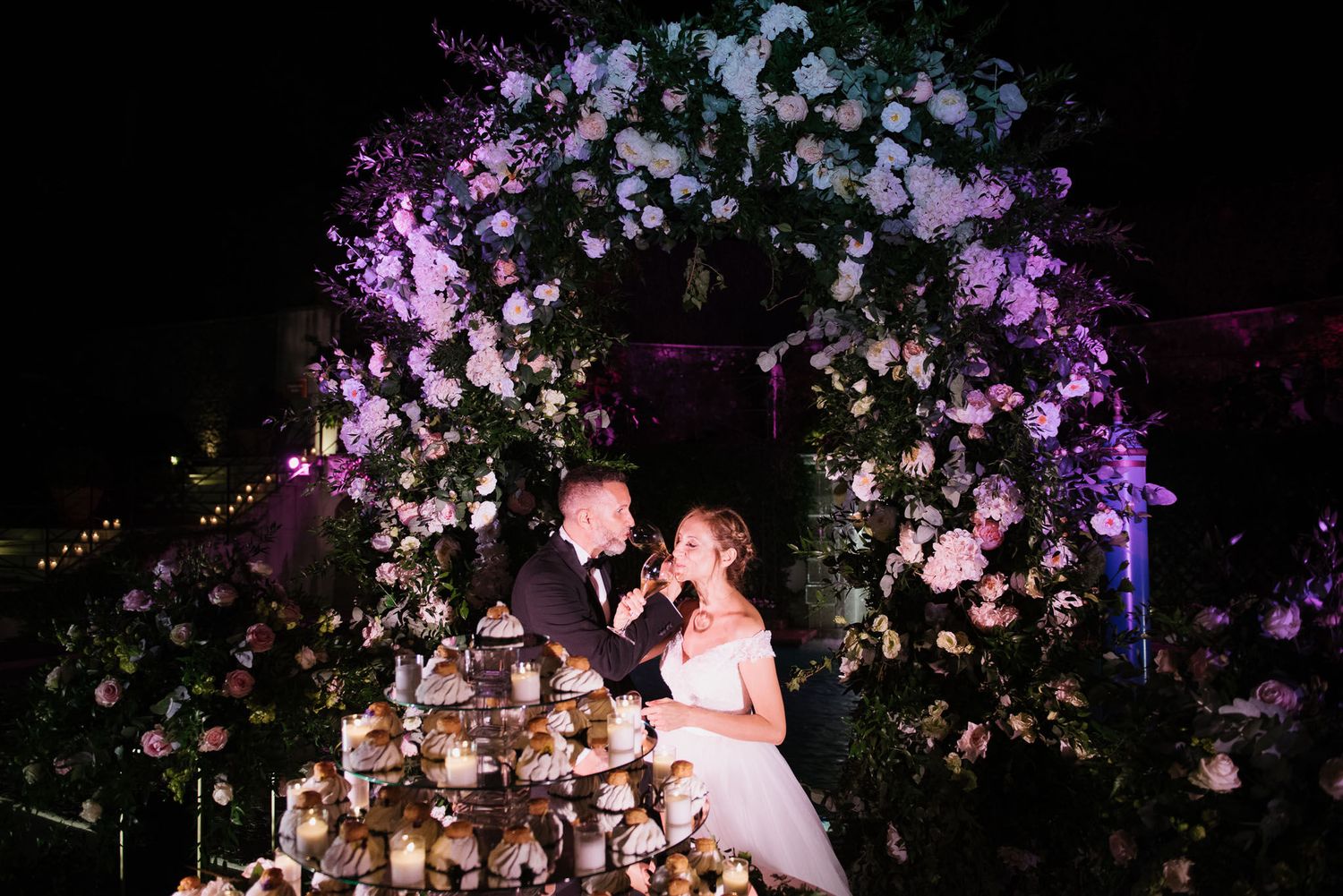 An elegant wedding dessert display under a floral arch with dramatic purple lighting.