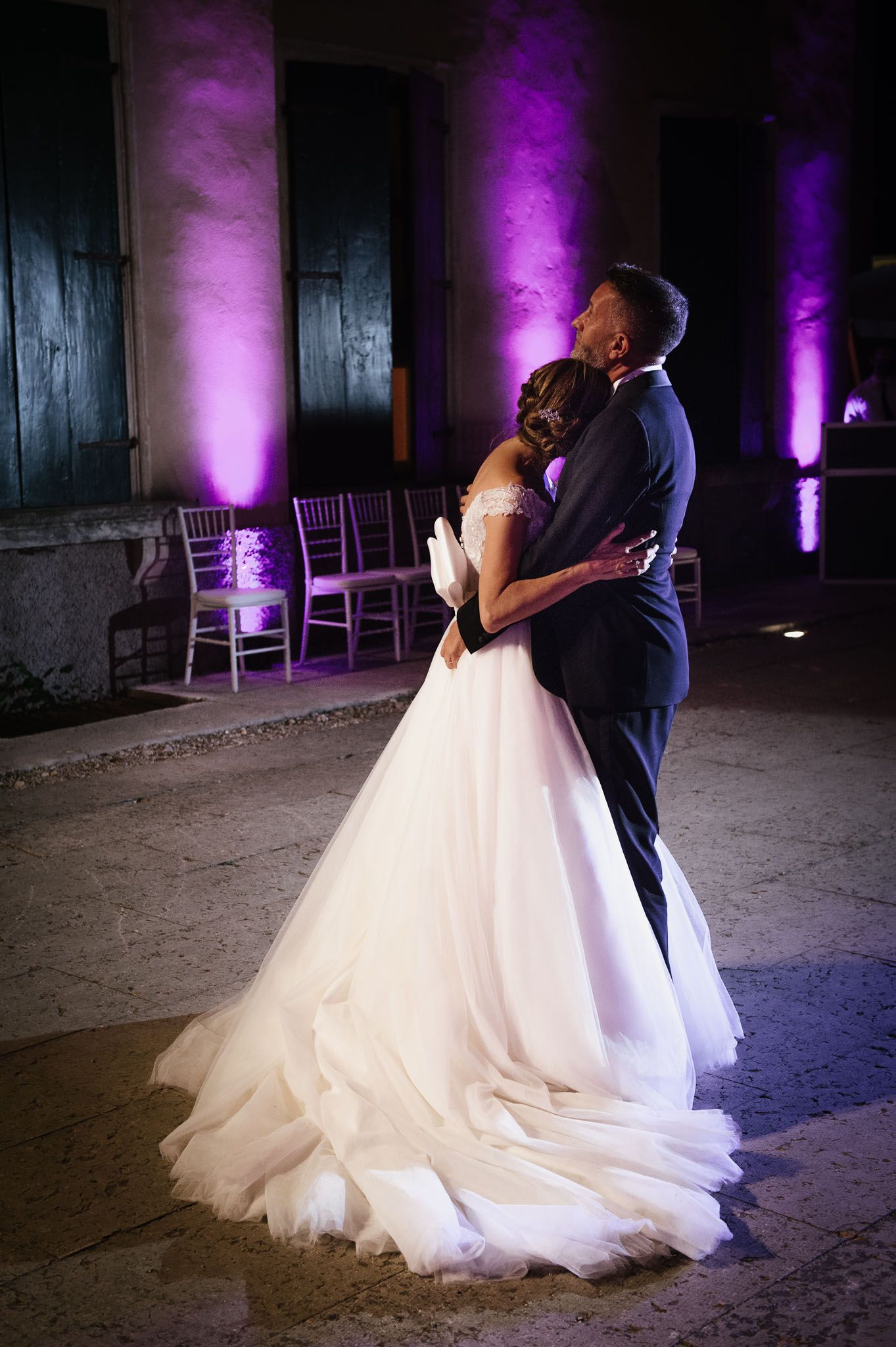 Newlyweds share a tender dance moment under purple-lit columns.