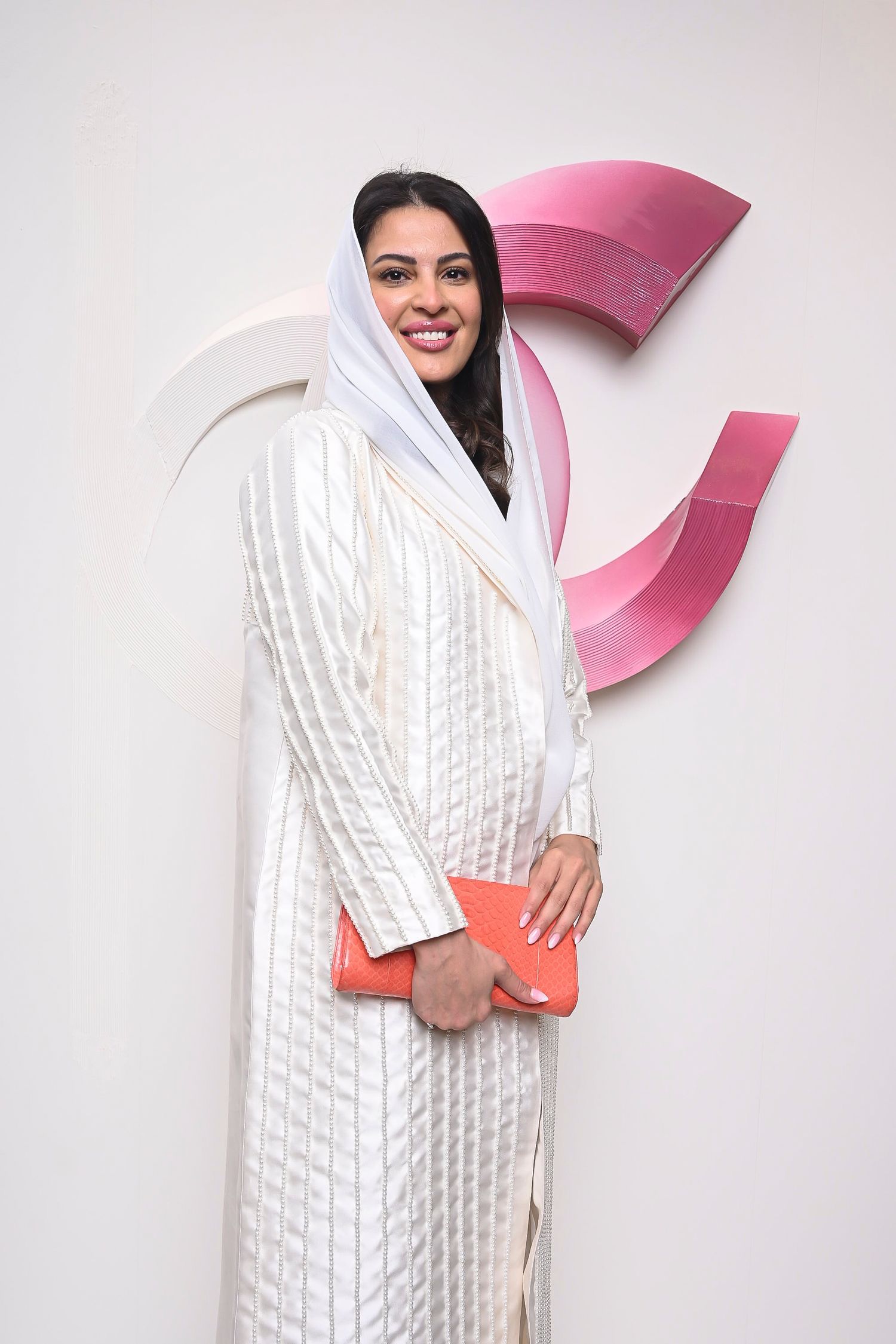 Woman in white traditional dress holds coral clutch against pink and white branded wall.