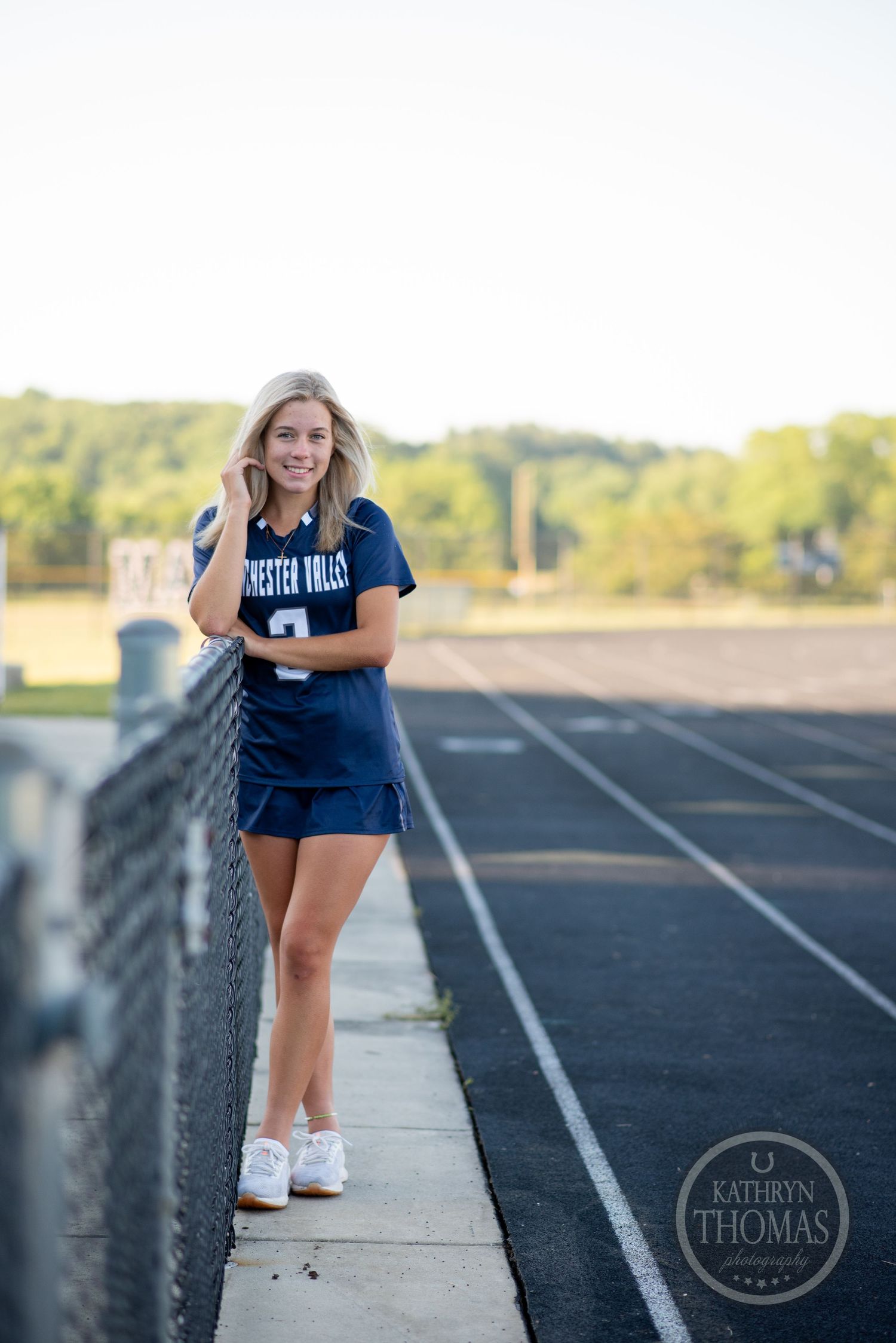Student athlete poses along track fence wearing navy uniform and white sneakers.
