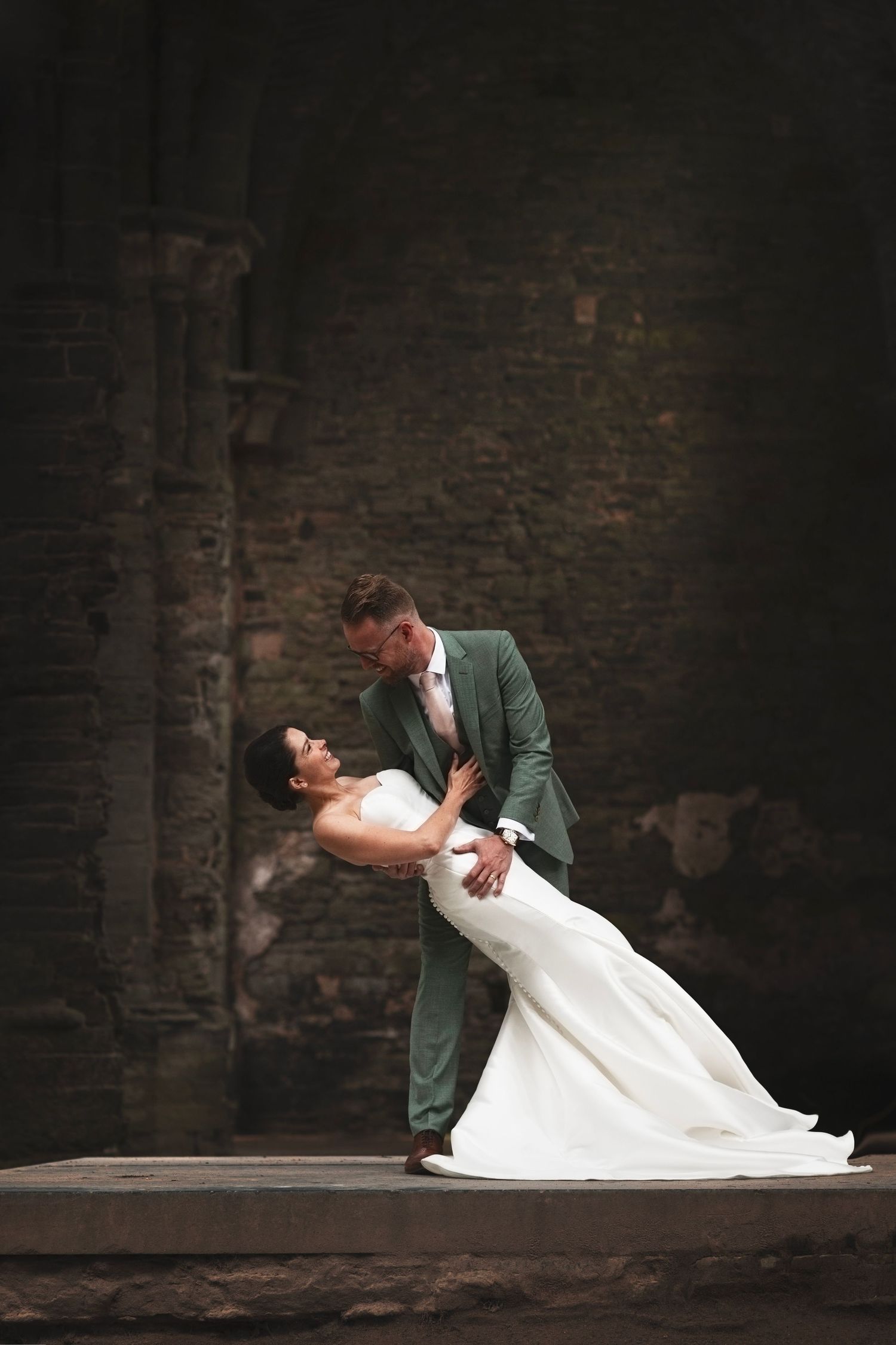 Dramatic wedding pose with couple performing dance dip against dark stone wall.