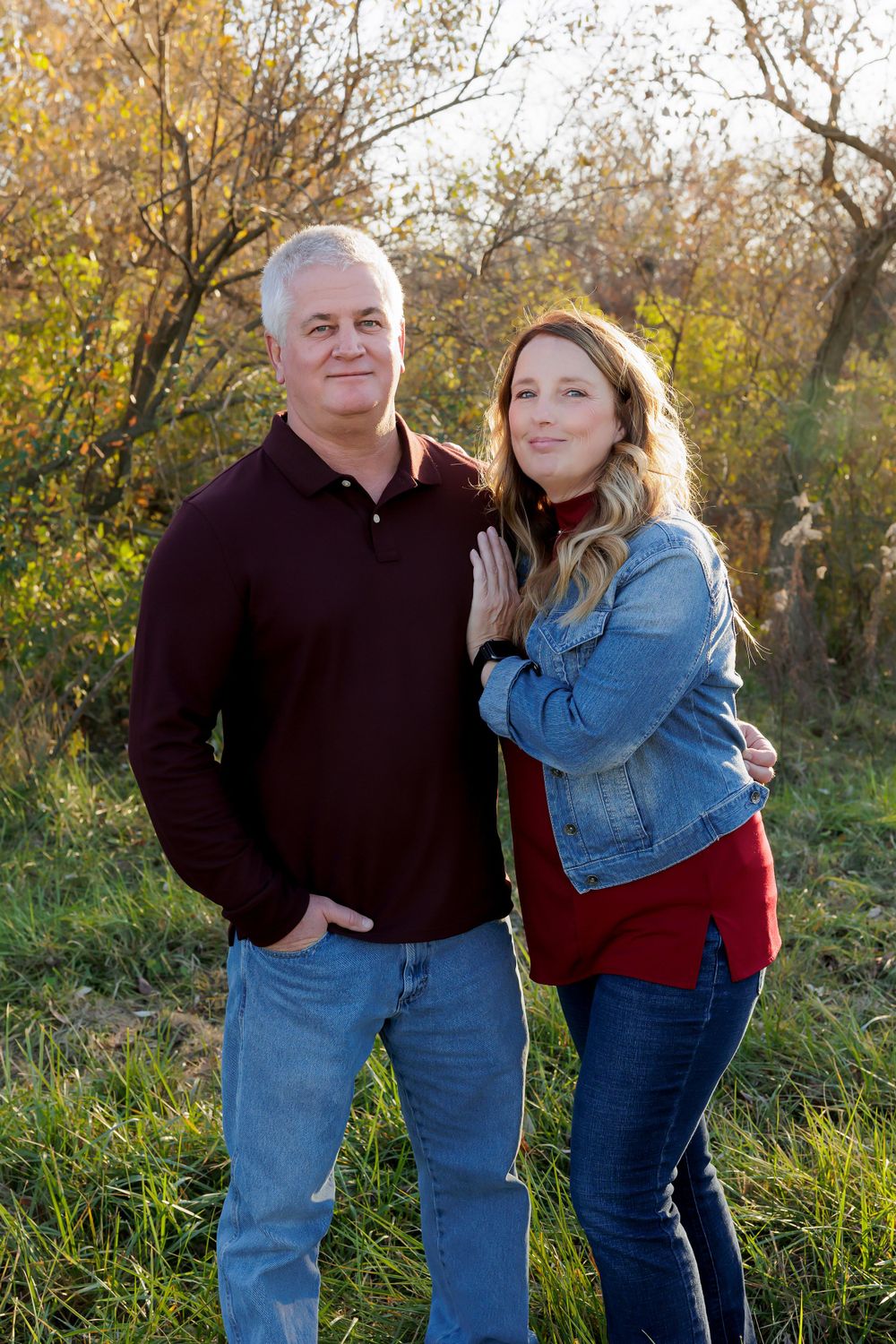 Two people stand close together for a casual autumn portrait with fall foliage in the background.