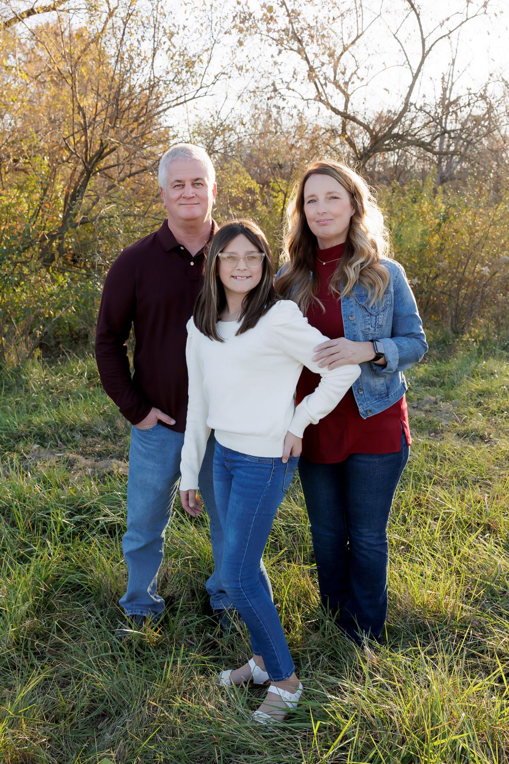 Three people in casual wear pose together for a family photo outdoors during autumn.