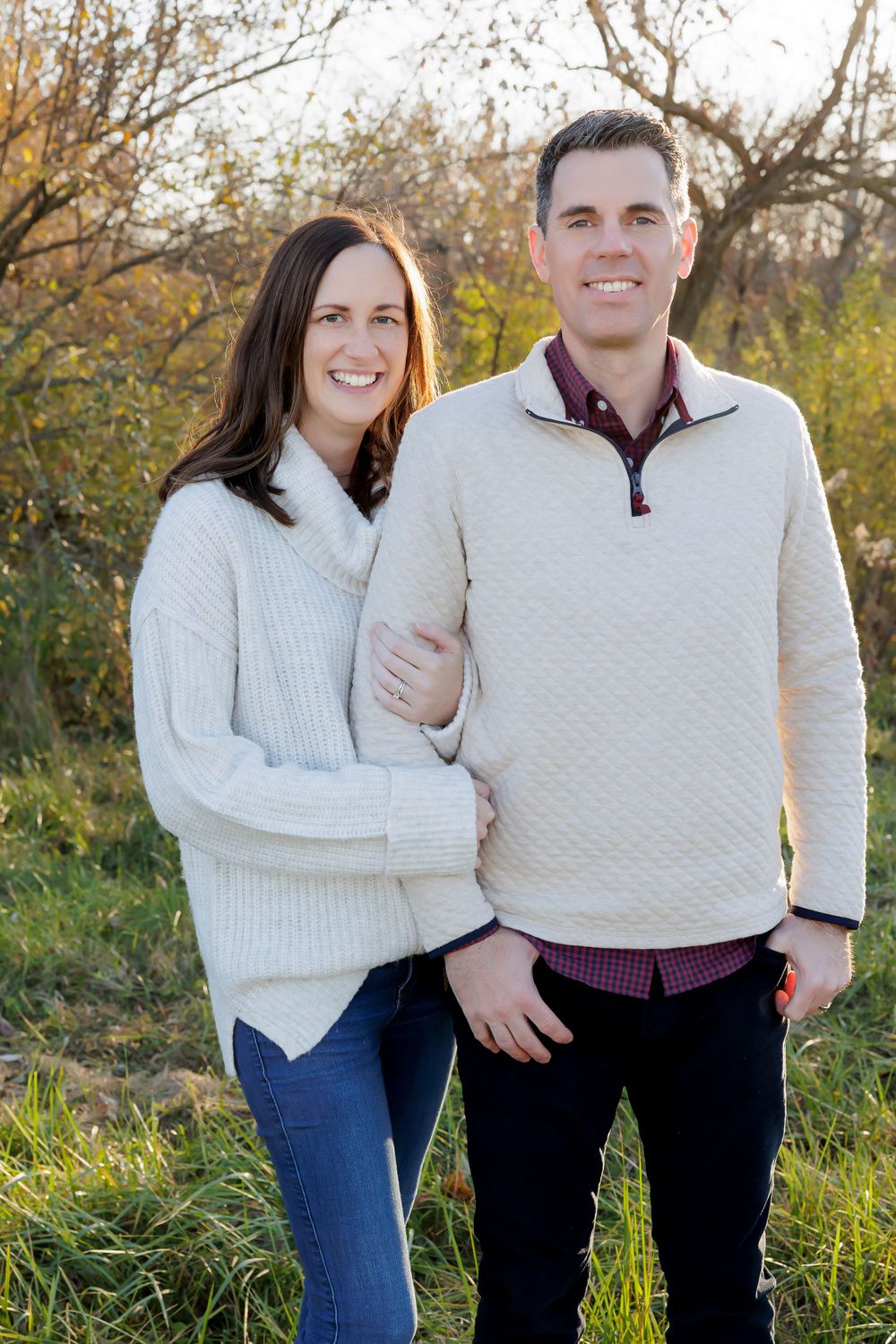 A couple wearing cozy sweaters smiles for a portrait in front of autumn trees and foliage.