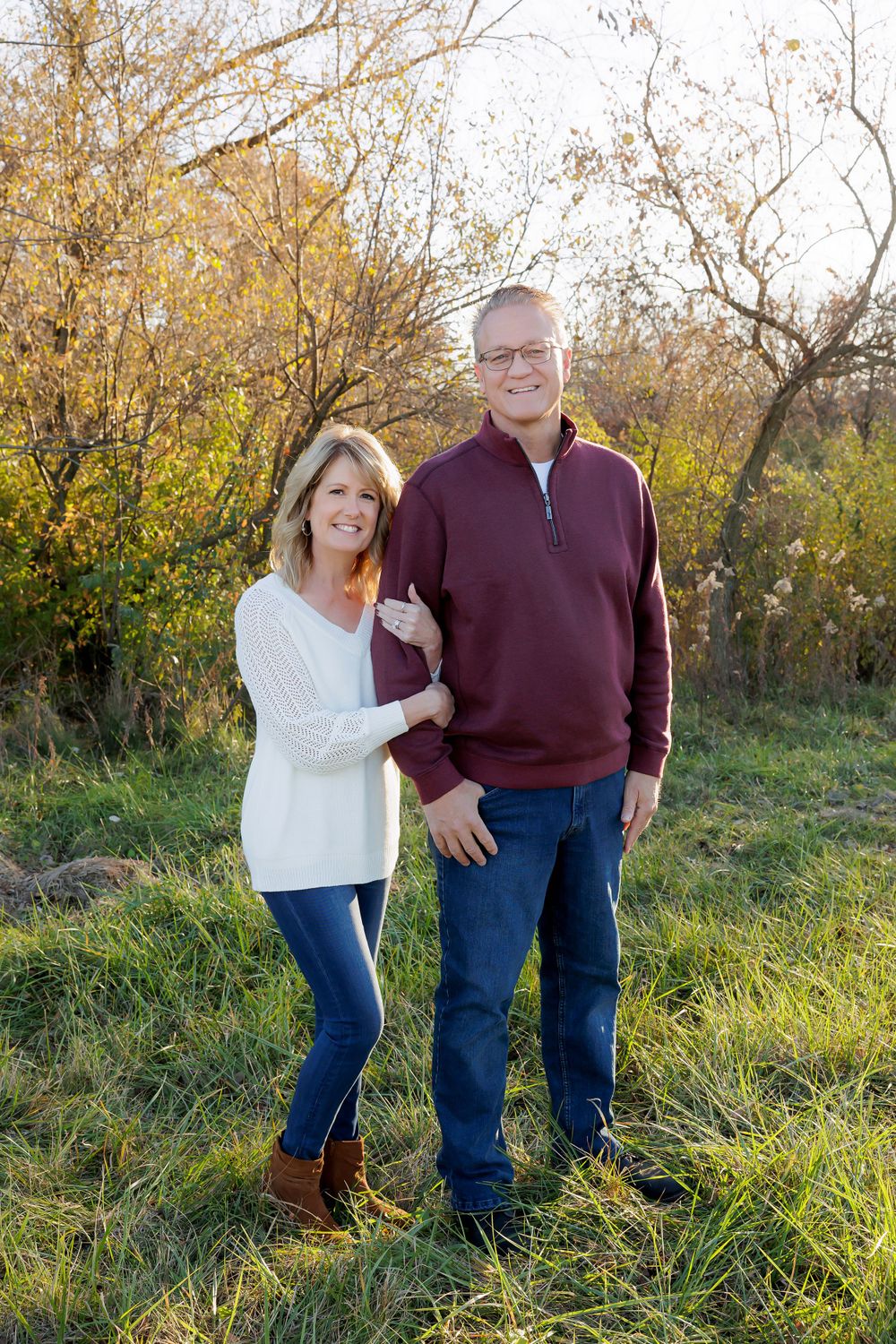 A couple in casual sweaters poses for a fall portrait in a natural outdoor setting with autumn foliage.