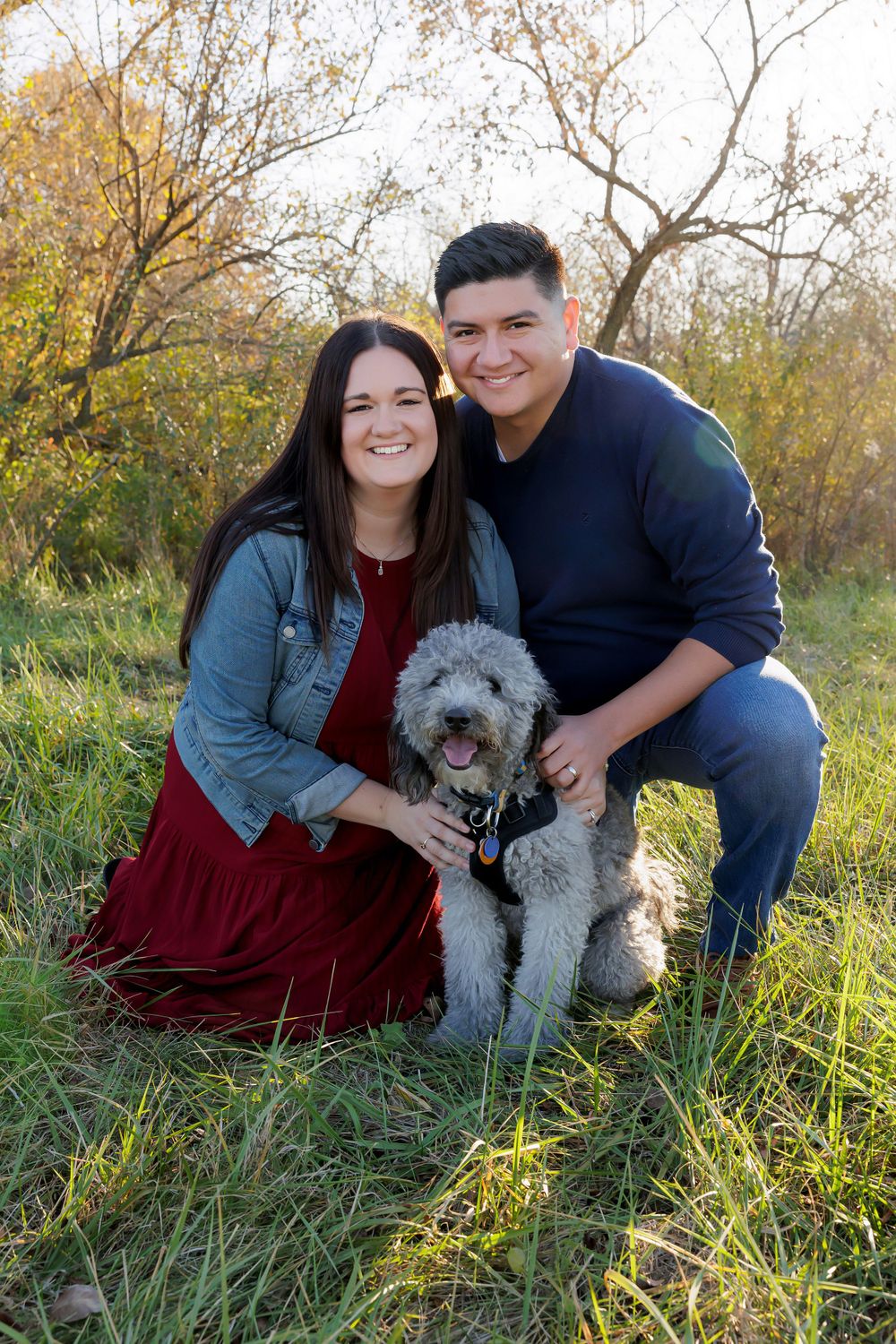 Couple sits with their fluffy grey dog in an outdoor autumn setting.
