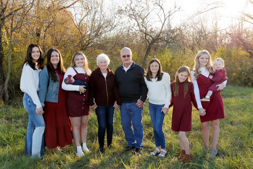 A group of eight people poses for a family portrait outdoors with fall foliage.