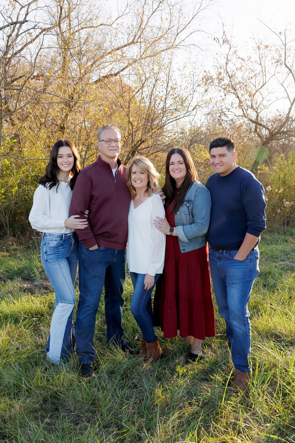 Family group of five poses together in casual attire against autumn foliage.