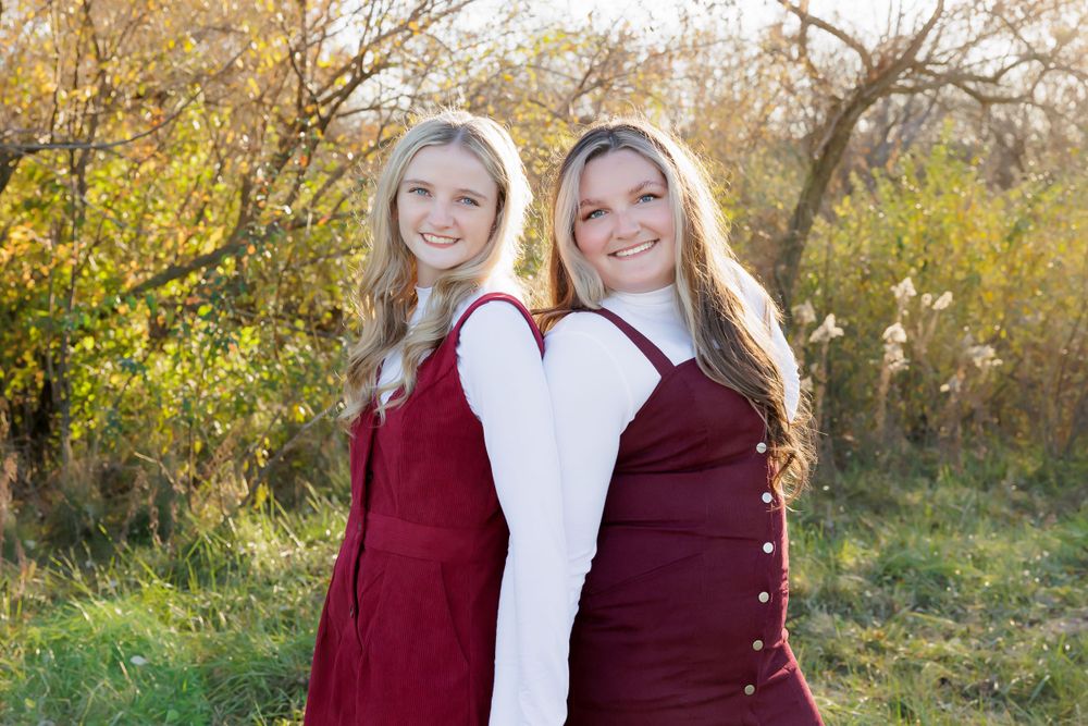 Two women in burgundy dresses pose back-to-back in an autumn outdoor setting.