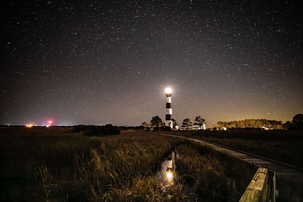Starlit night sky over a lighthouse and coastal landscape.