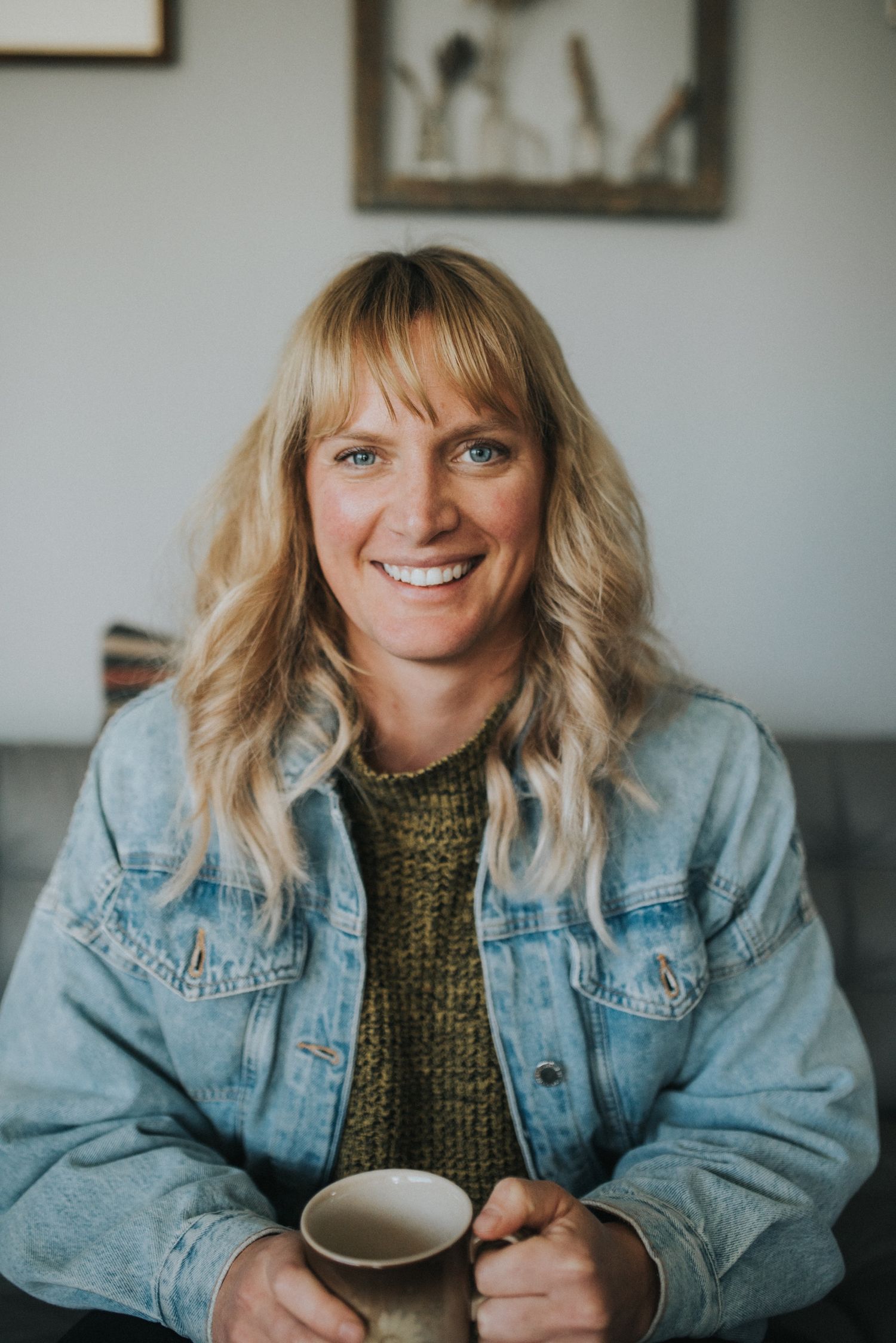 Casual indoor portrait with denim jacket and coffee cup against white wall.