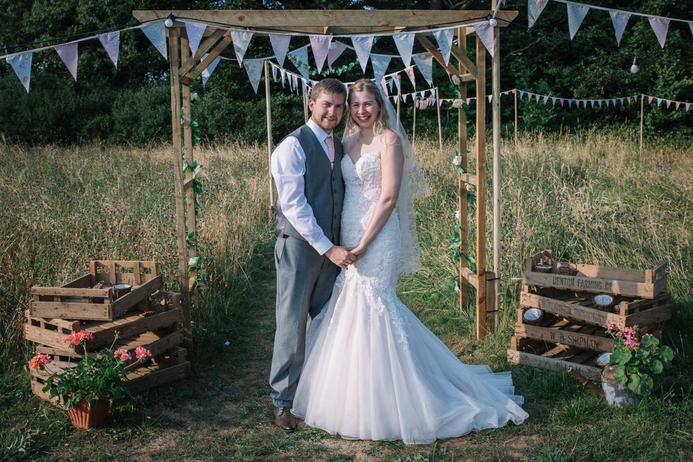 Couple stands under a wooden arch with bunting decorations and rustic crates.
