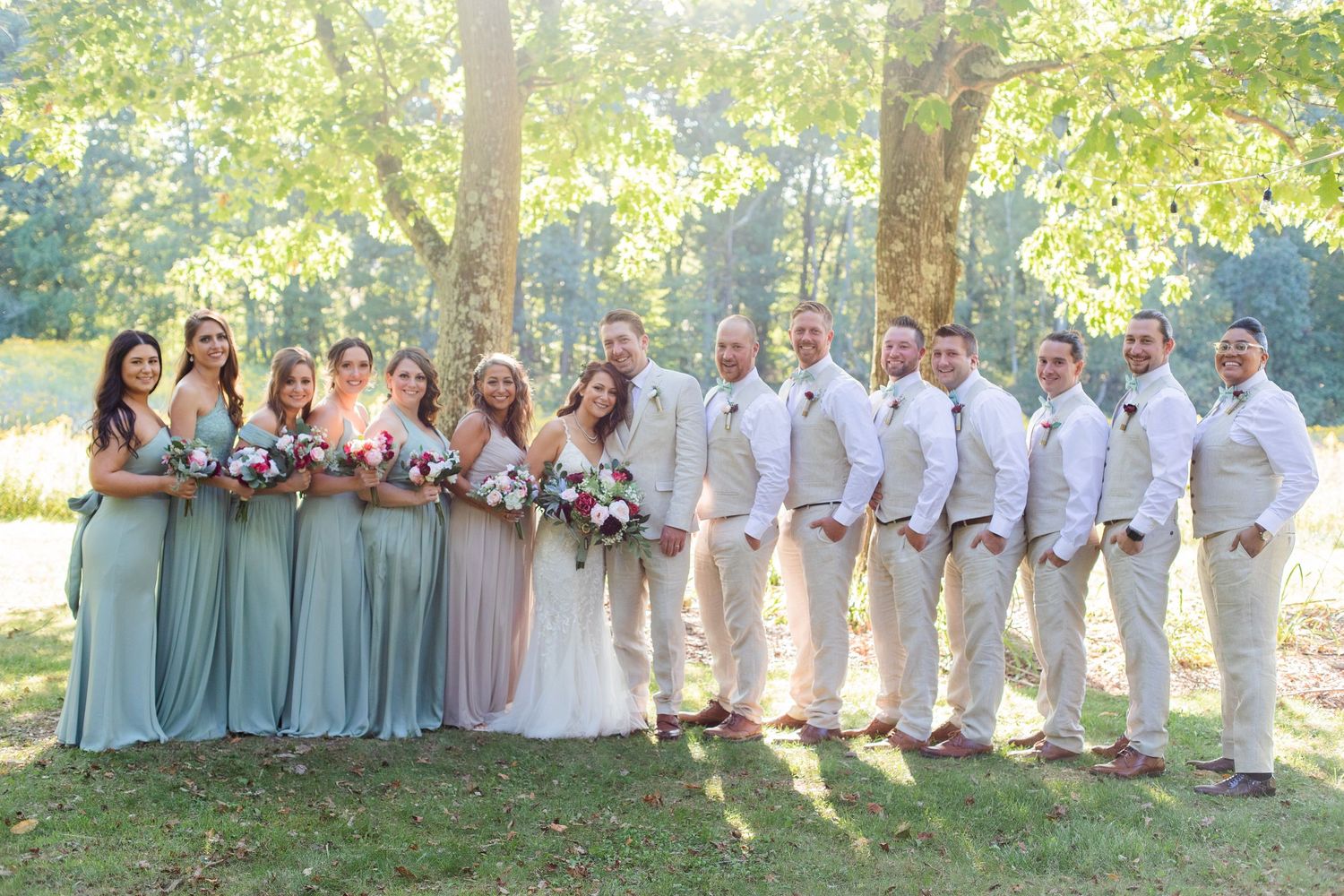Wedding party in sage green and white attire poses outdoors under tree canopy.