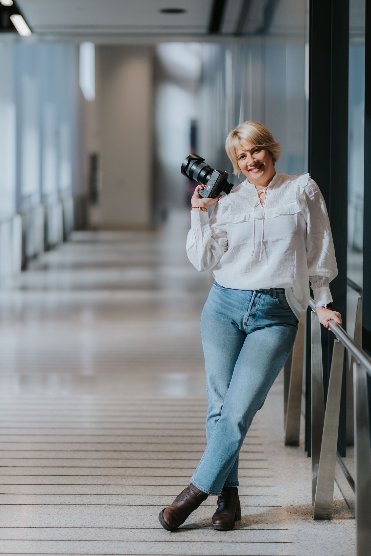 A professional photographer with camera poses casually in a modern corridor with natural light.