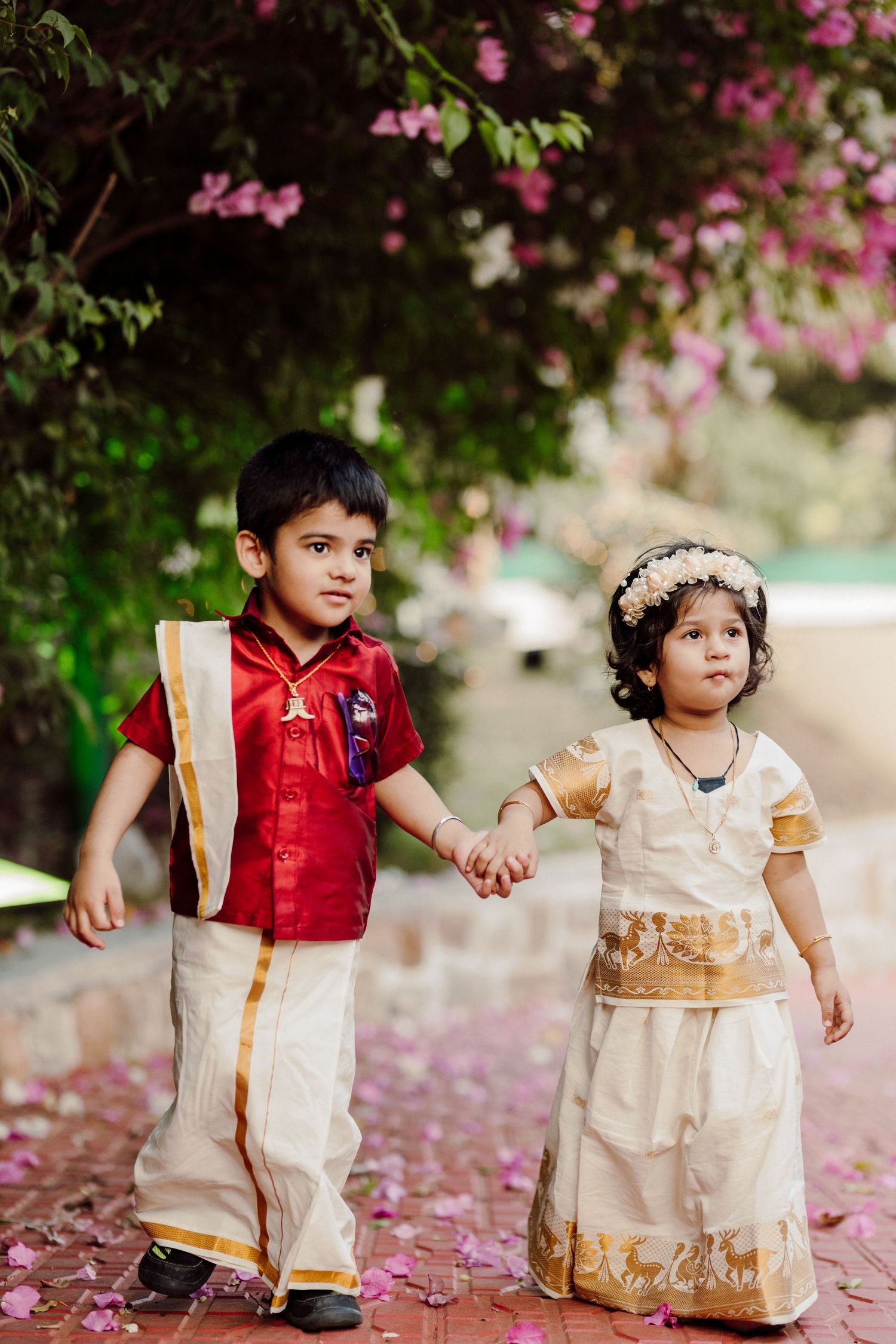 Two children in traditional Indian outfits playing on brick pathway under pink flowering trees.
