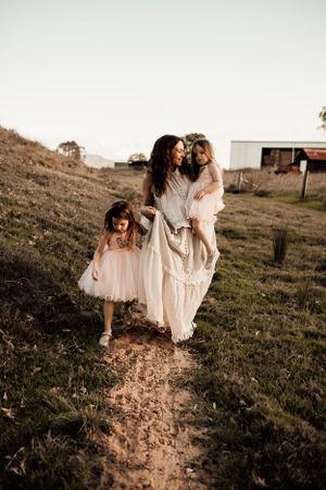 Three figures in flowing white dresses walk along a rural dirt path at sunset.