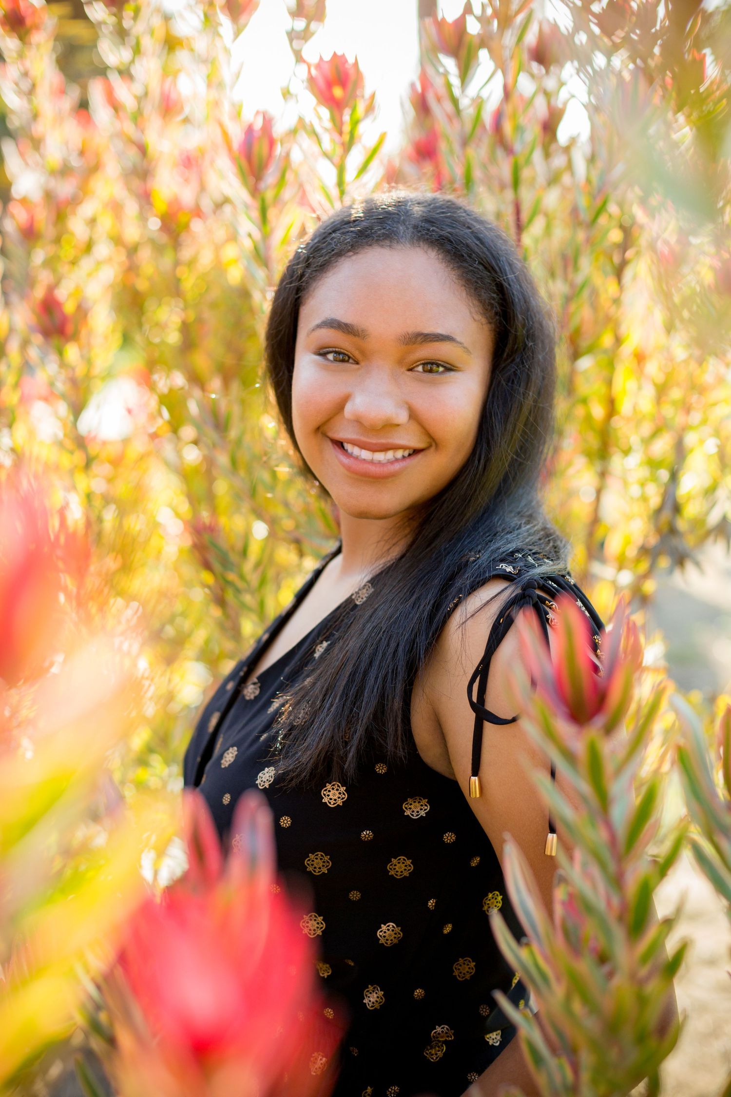 A smiling woman in a black polka dot top poses among red flowering plants on a sunny day.