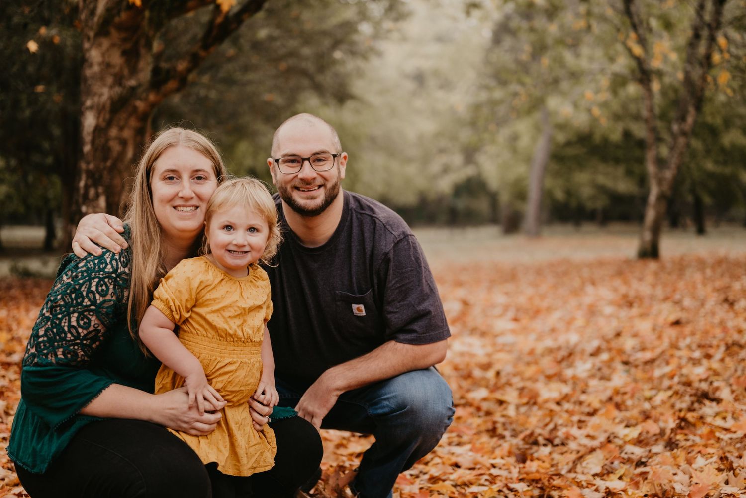 Family of three poses together among fallen autumn leaves in a park setting.