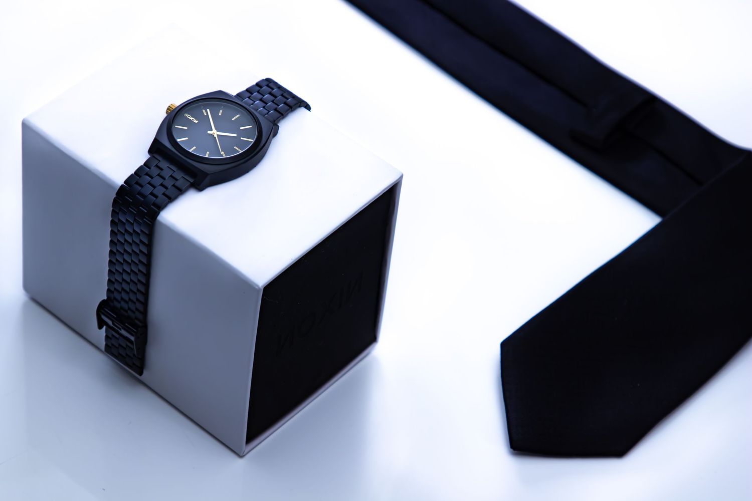 Minimalist product photography of a dark watch and tie on white geometric shapes.