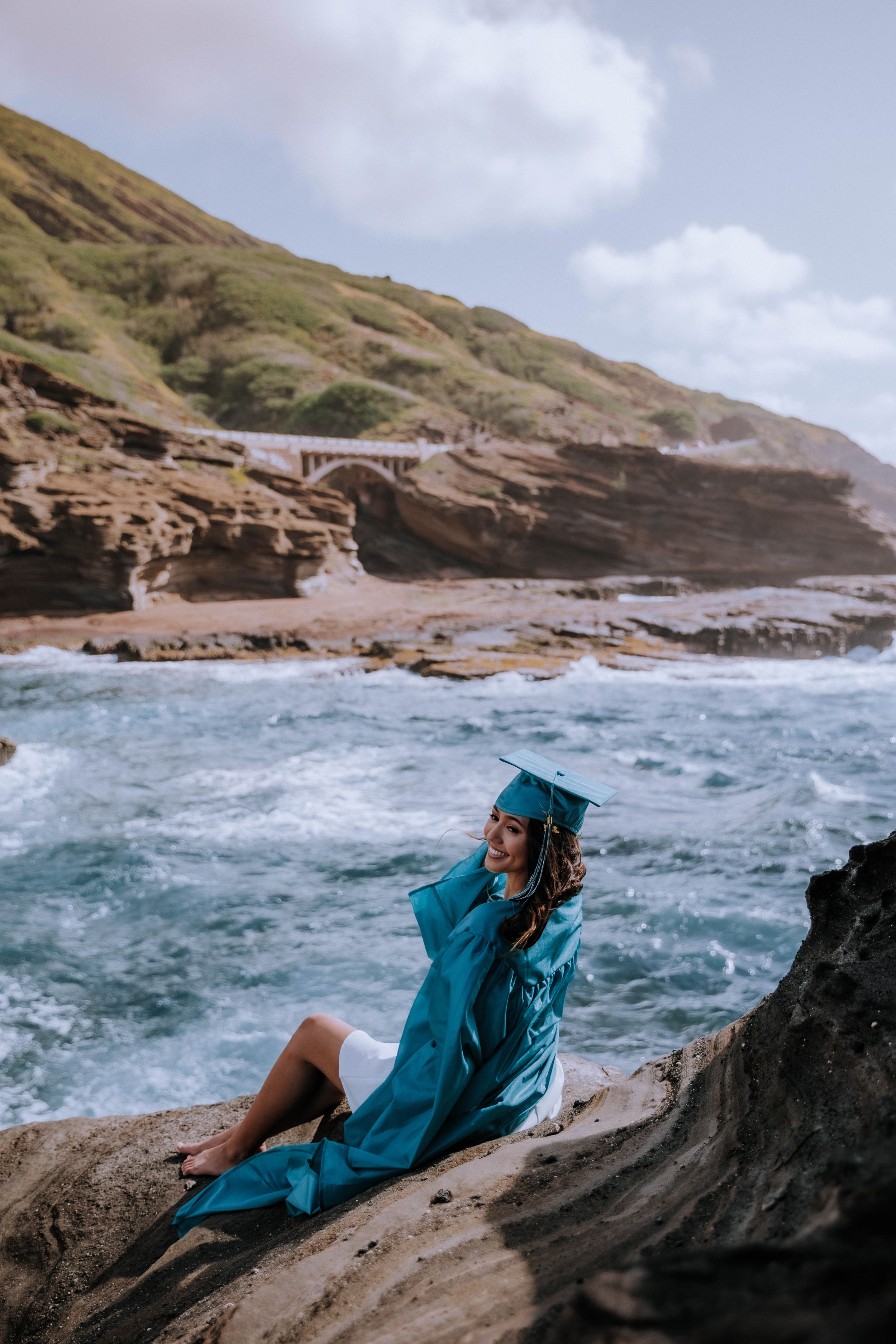 A person in a teal dress sits on rocky coastal cliffs overlooking dramatic ocean waves.