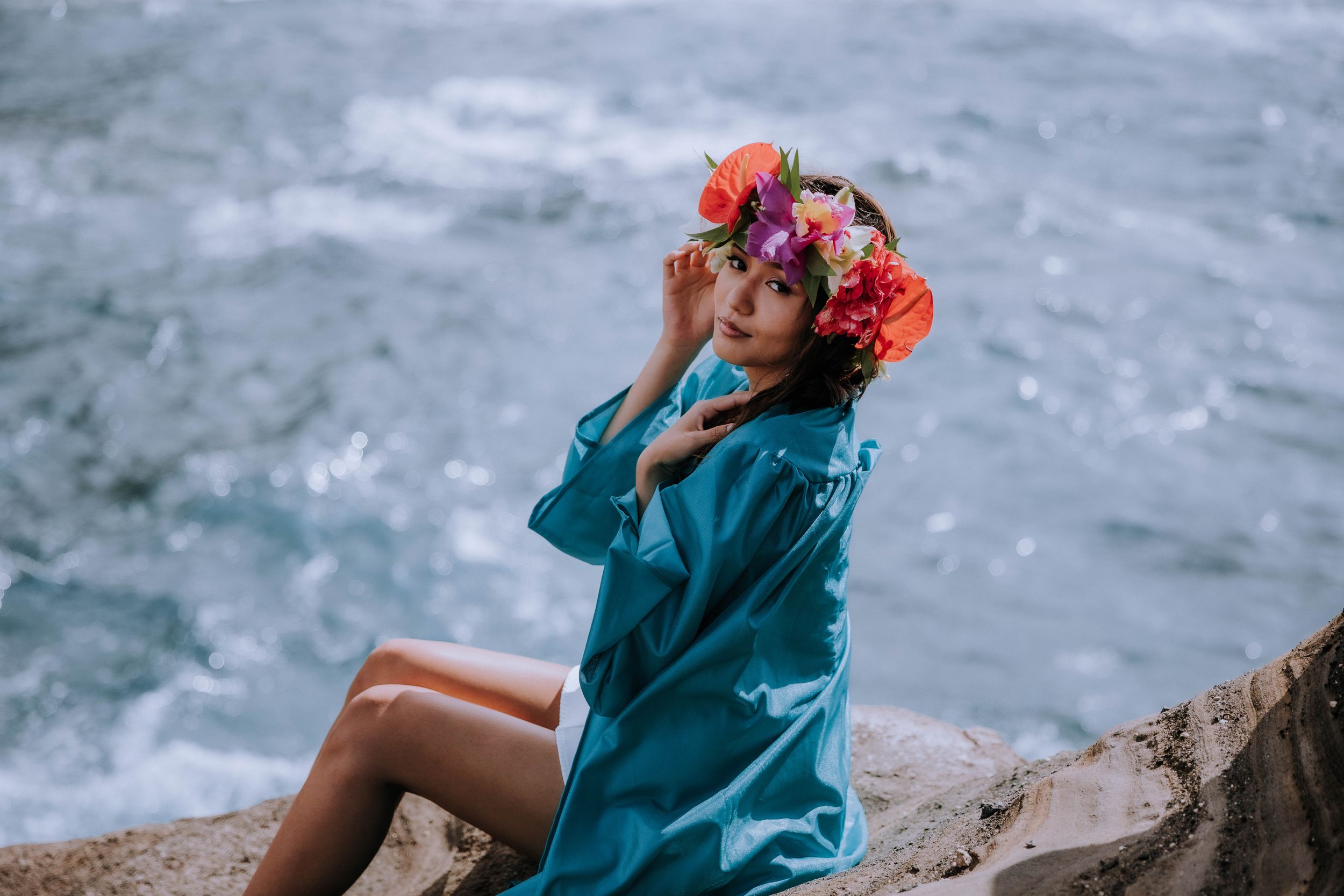 A dreamy coastal photoshoot featuring someone in turquoise clothing with pink flowers against ocean cliffs.