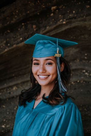 A graduation portrait featuring someone in teal academic regalia smiling against a dark background.