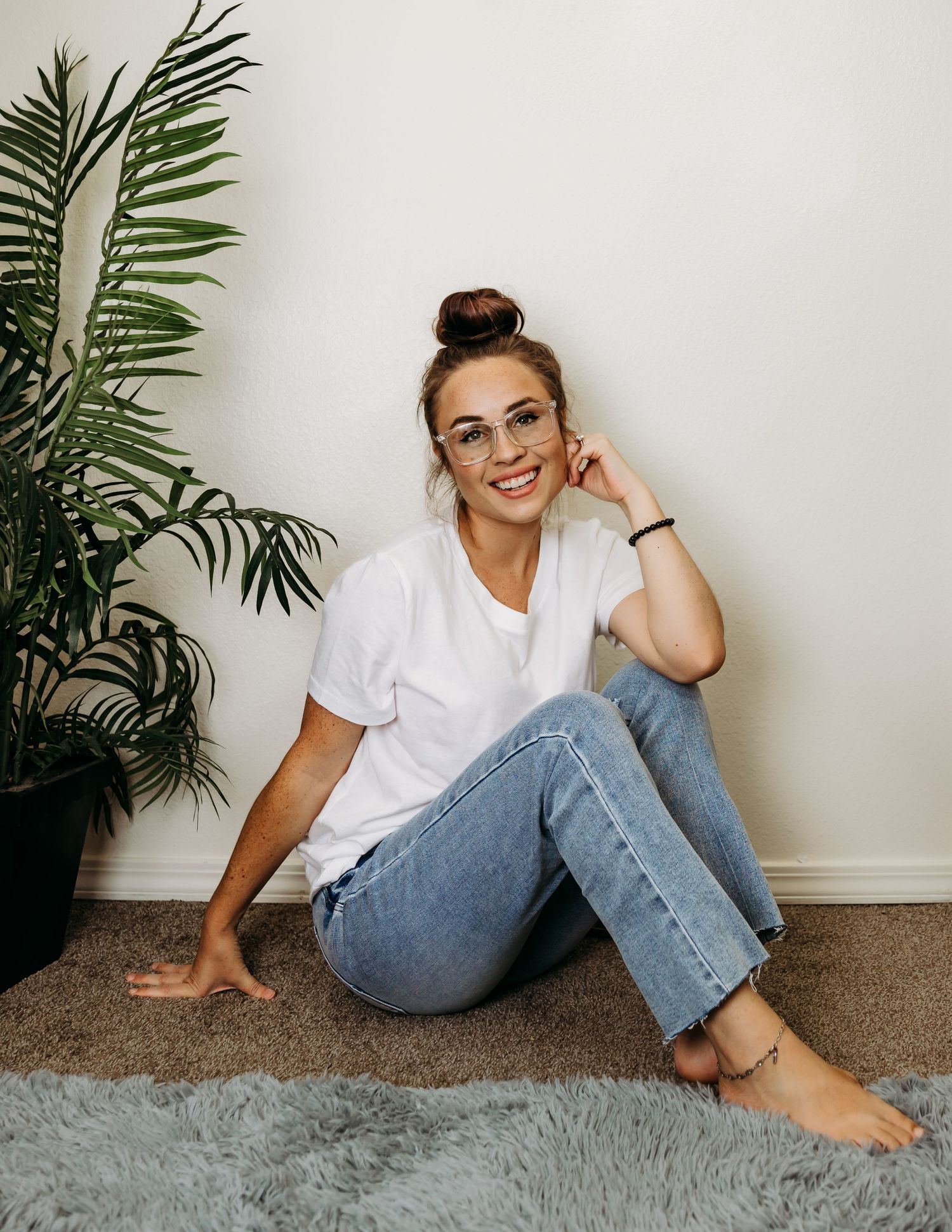 Smiling woman with glasses in white tee and jeans sitting on rug near plant.
