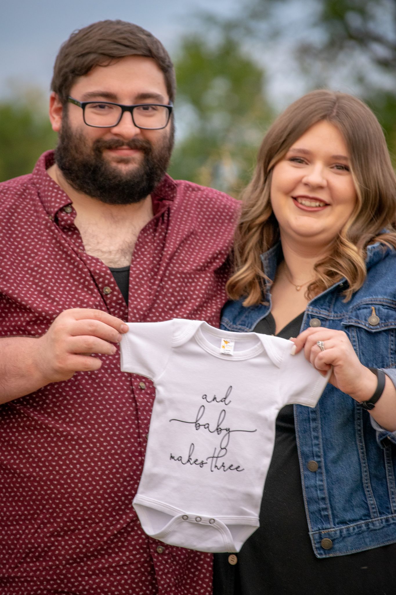 Pregnancy Announcement Session. Mom and Dad hold a Onesie that reads "And baby makes three" smiling at the camera