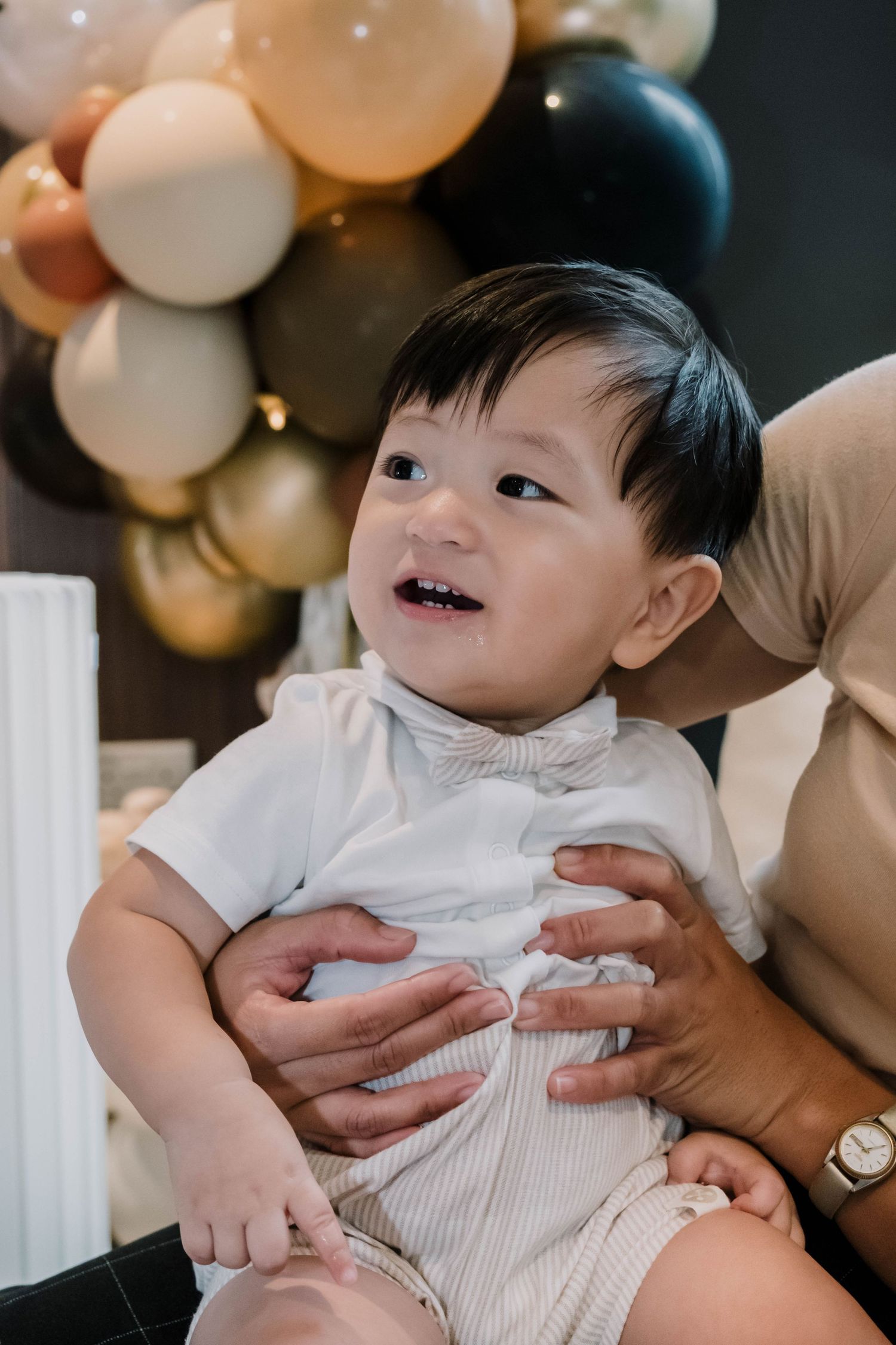 Baby in white outfit smiling with balloon decorations in background.