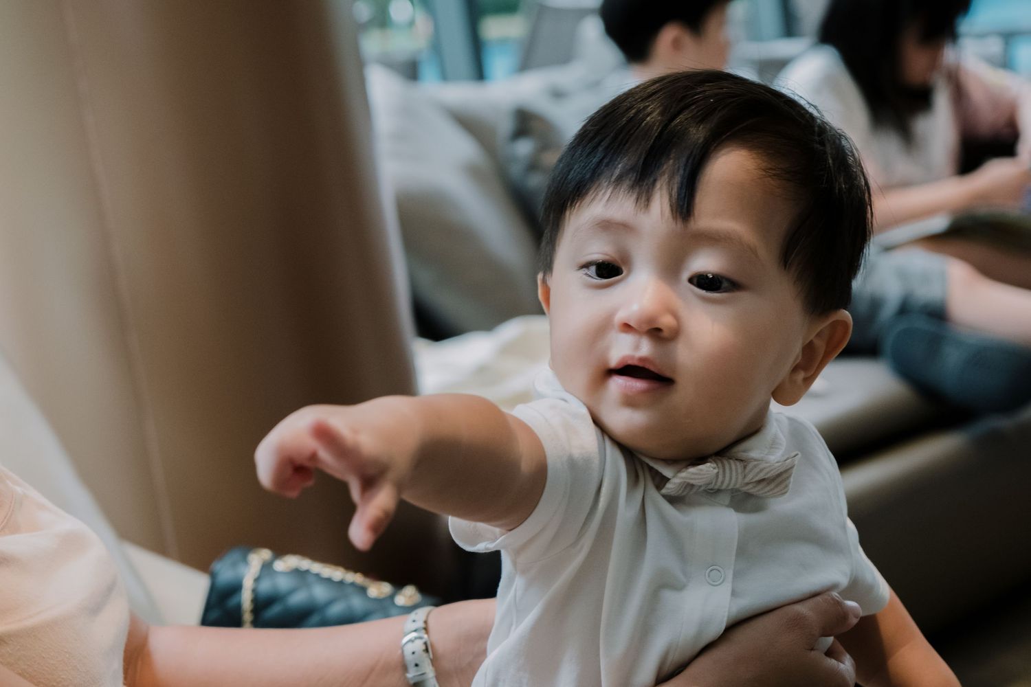 Toddler in white shirt pointing while sitting indoors.