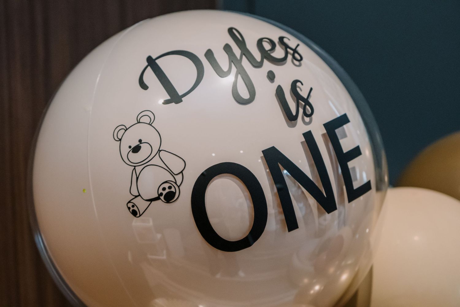 White balloon with 'Dylan is ONE' text and teddy bear illustration.