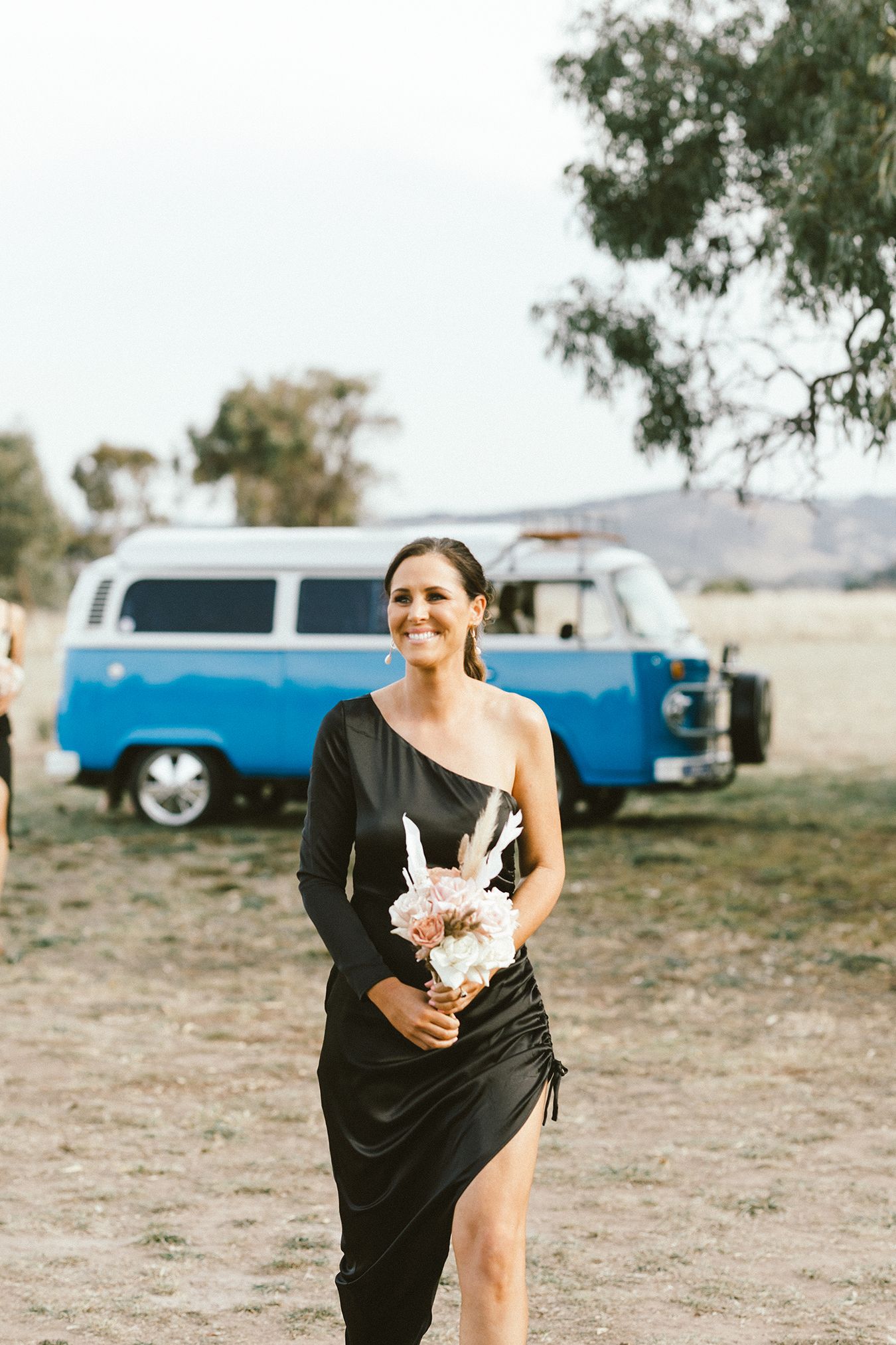 Bridesmaid in black dress carrying bouquet with blue vintage VW van in background.