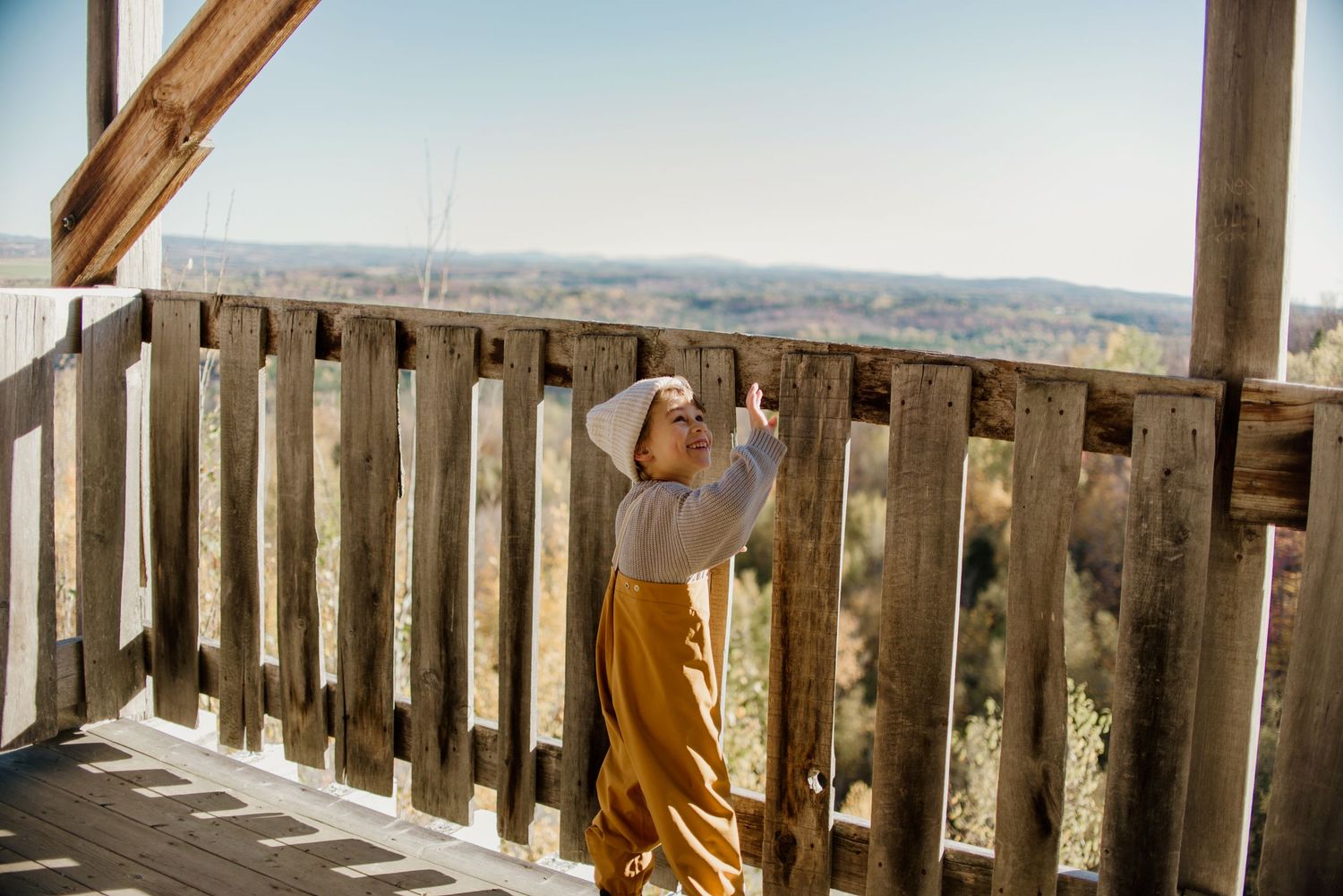 Person wearing yellow overalls and white hat standing at rustic wooden deck railing overlooking scenic landscape.