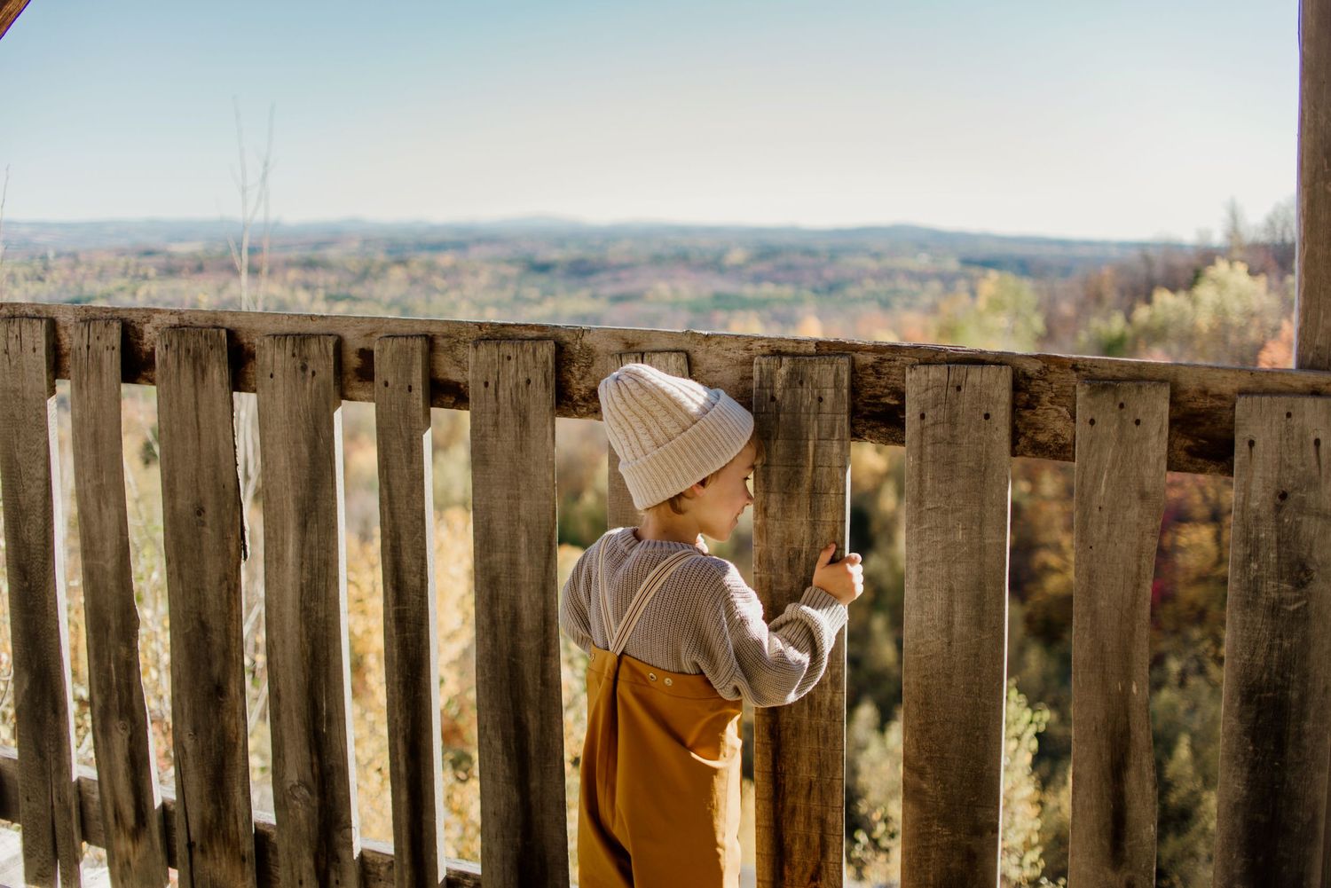 Figure in mustard overalls and beanie looking out over countryside from wooden observation deck.