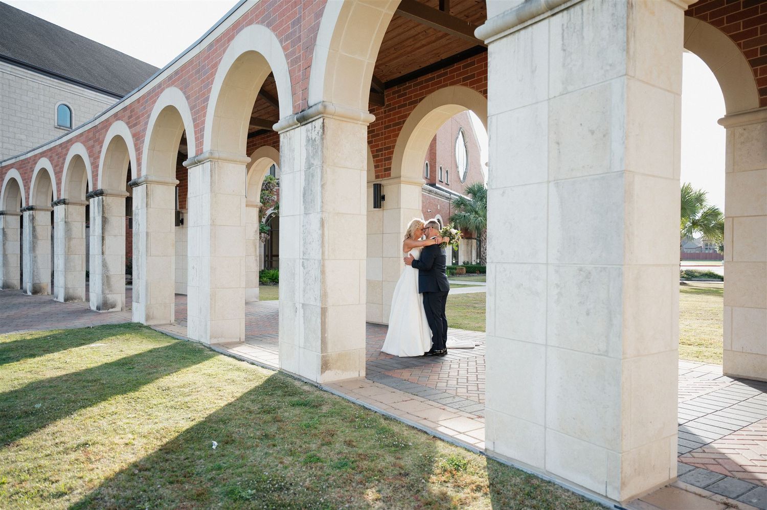 A wedding couple embraces under a beautiful colonnade with brick and stone architecture.